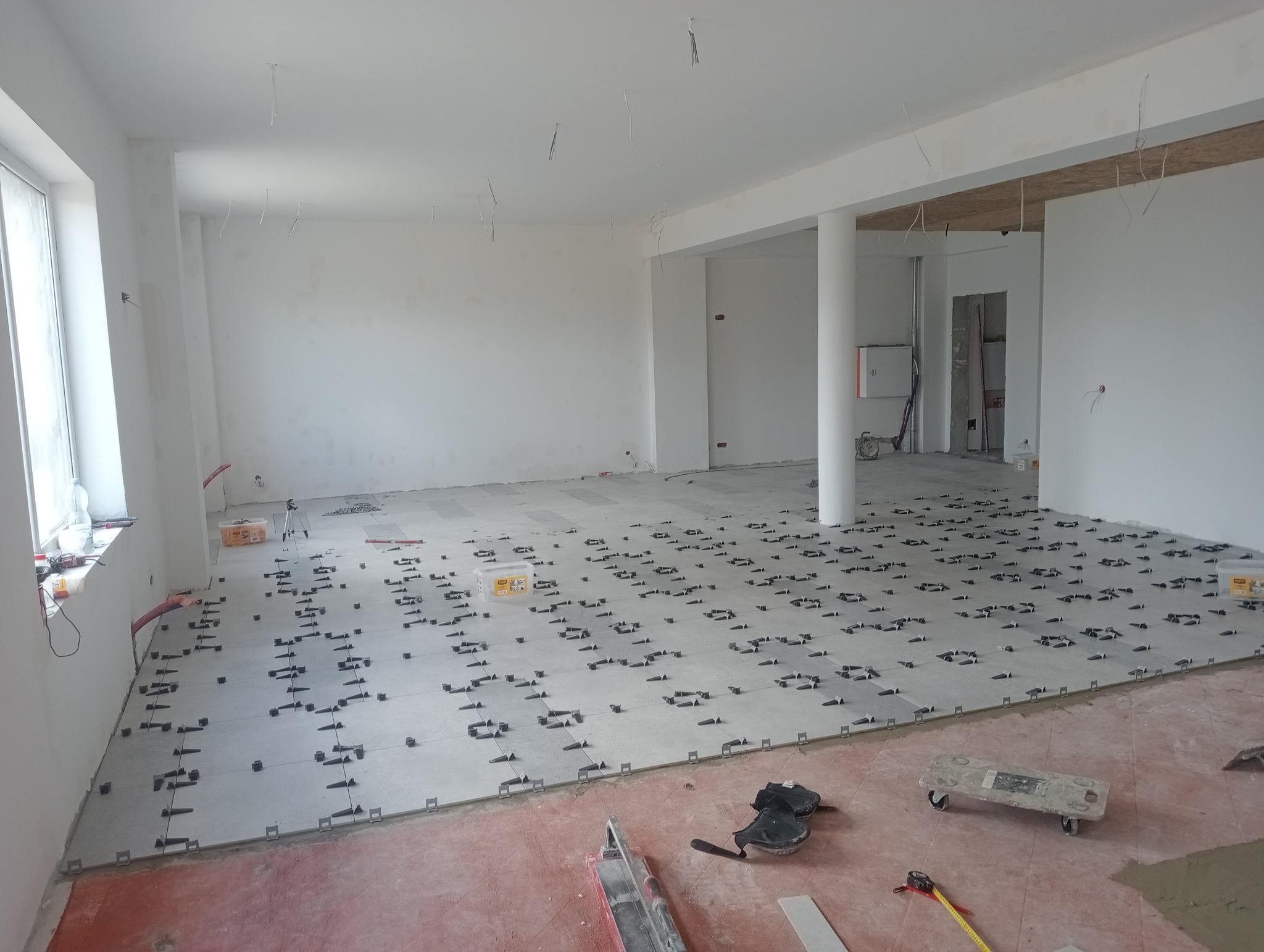 Jasne wnętrze w trakcie układania płytek podłogowych z użyciem klipsów poziomujących. Widoczne narzędzia i akcesoria glazurnicze.