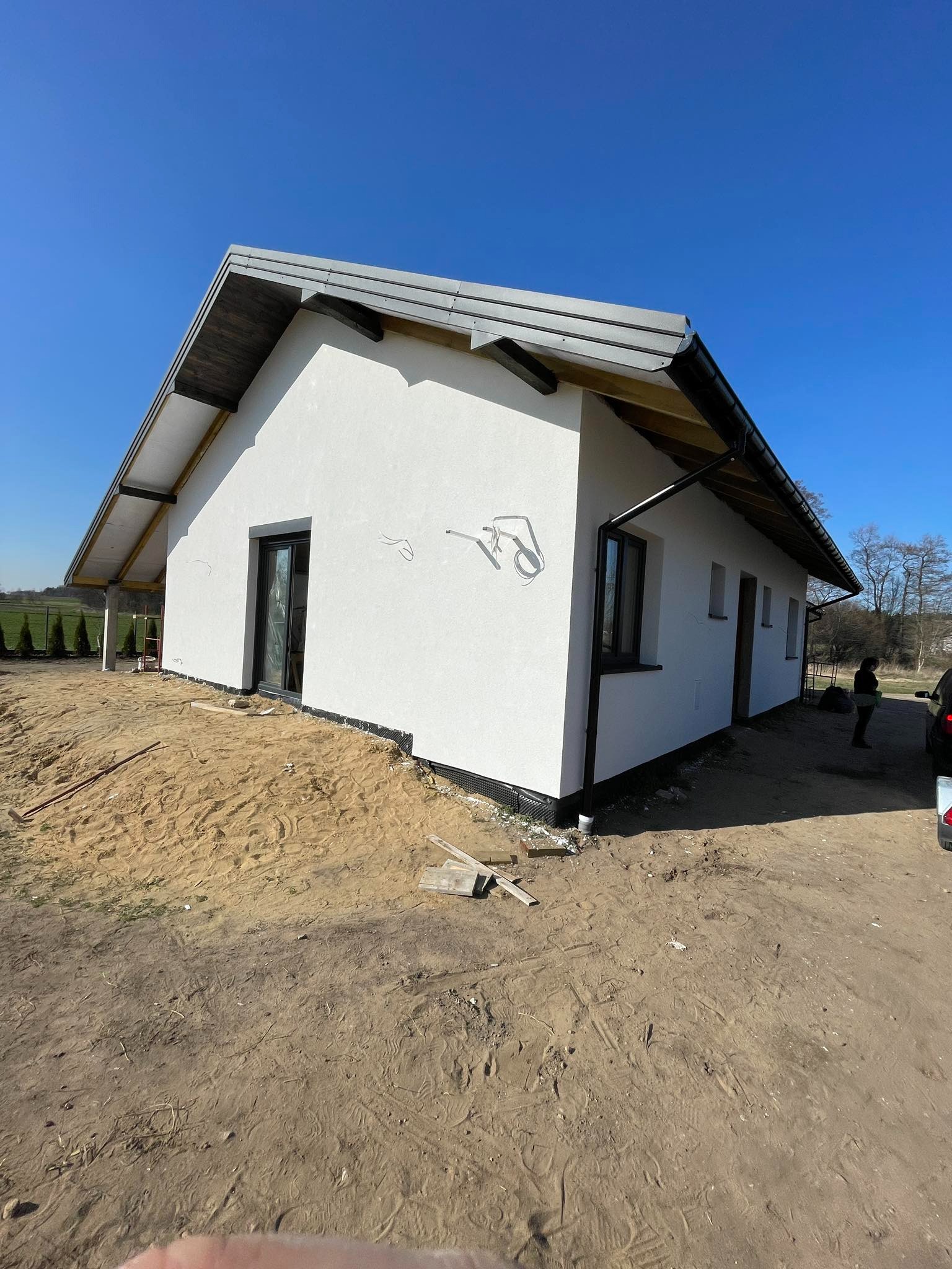 Biały dom jednorodzinny z ciemnymi oknami i dachem, w trakcie prac wykończeniowych elewacji. Widoczne ślady po ołówku na ścianie i piasek wokół budynku.