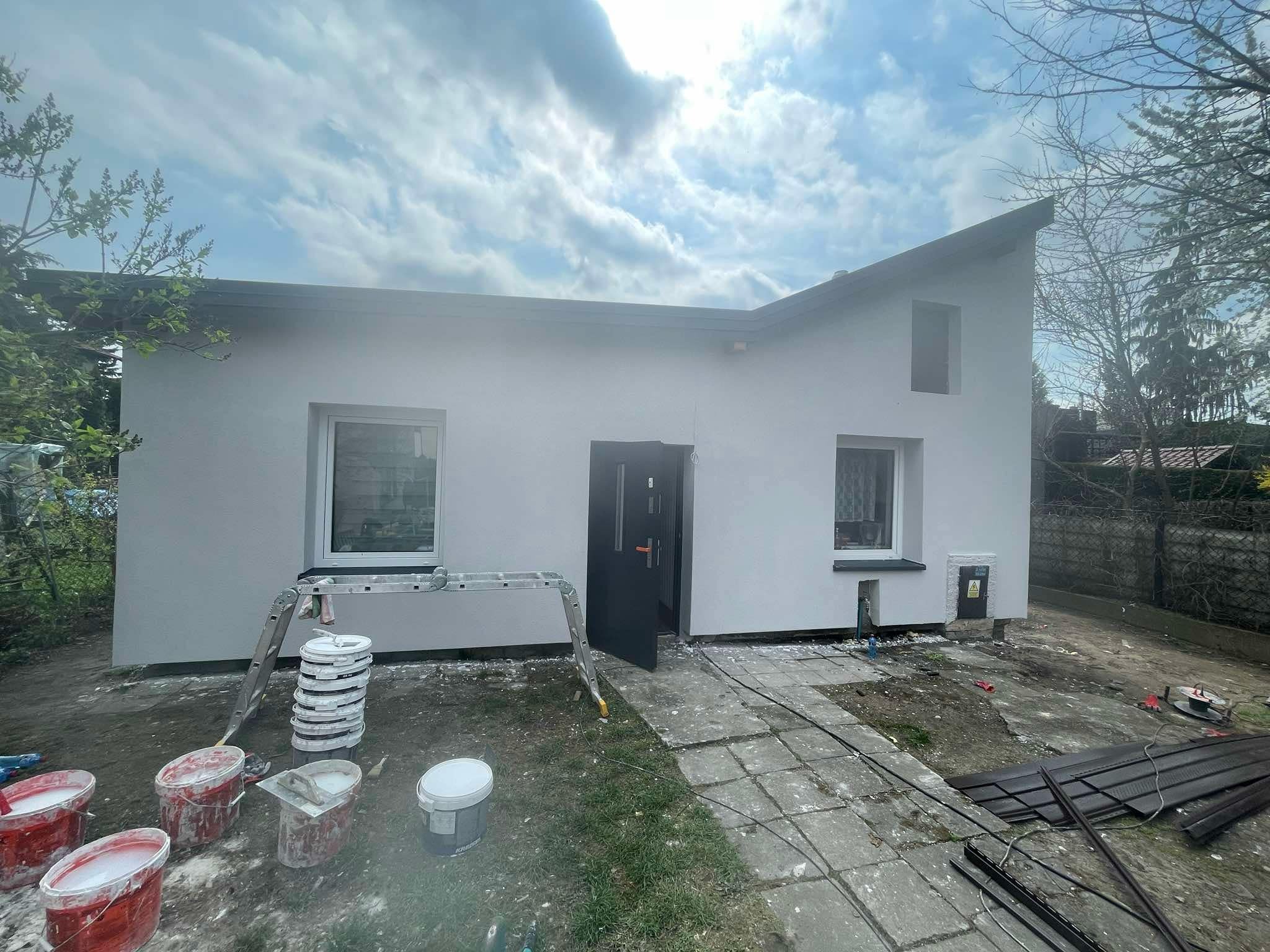 Dom jednorodzinny po malowaniu elewacji, widoczne drabiny, wiaderka z farbą i świeżo pomalowana fasada w odcieniach bieli i szarości. Ujęcie z poziomu gruntu.