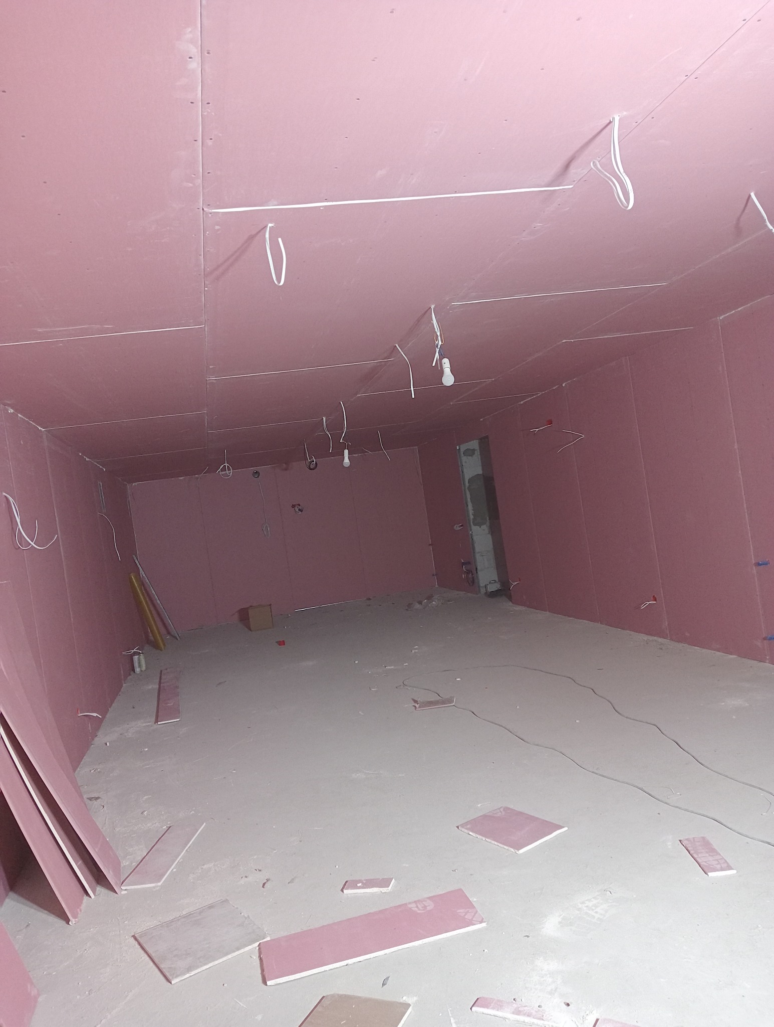 Różowe płyty na ścianach i suficie w surowym wnętrzu. Widoczne kable i fragmenty płyt na podłodze. W tle otwarte przejście do innego pomieszczenia.