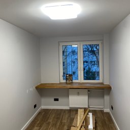 Labut Perfekt Remont - Jasne wnętrze po remoncie: świeżo pomalowane ściany, drewniany blat pod oknem, nowe panele podłogowe. Widoczne materiały budowlane.