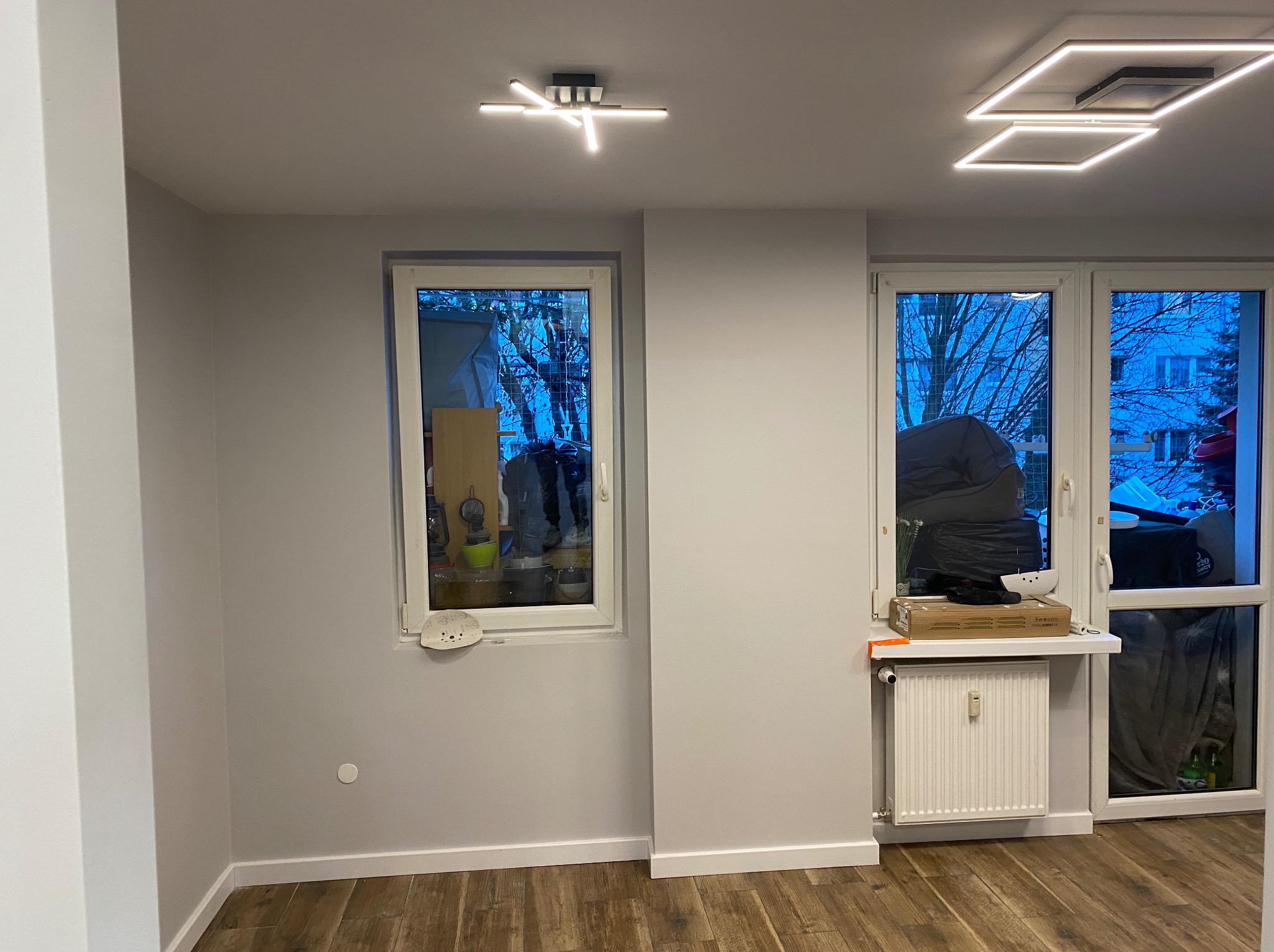 Jasne wnętrze po remoncie: szare ściany, drewnopodobne panele podłogowe, białe okna i grzejnik. Nowoczesne lampy sufitowe.