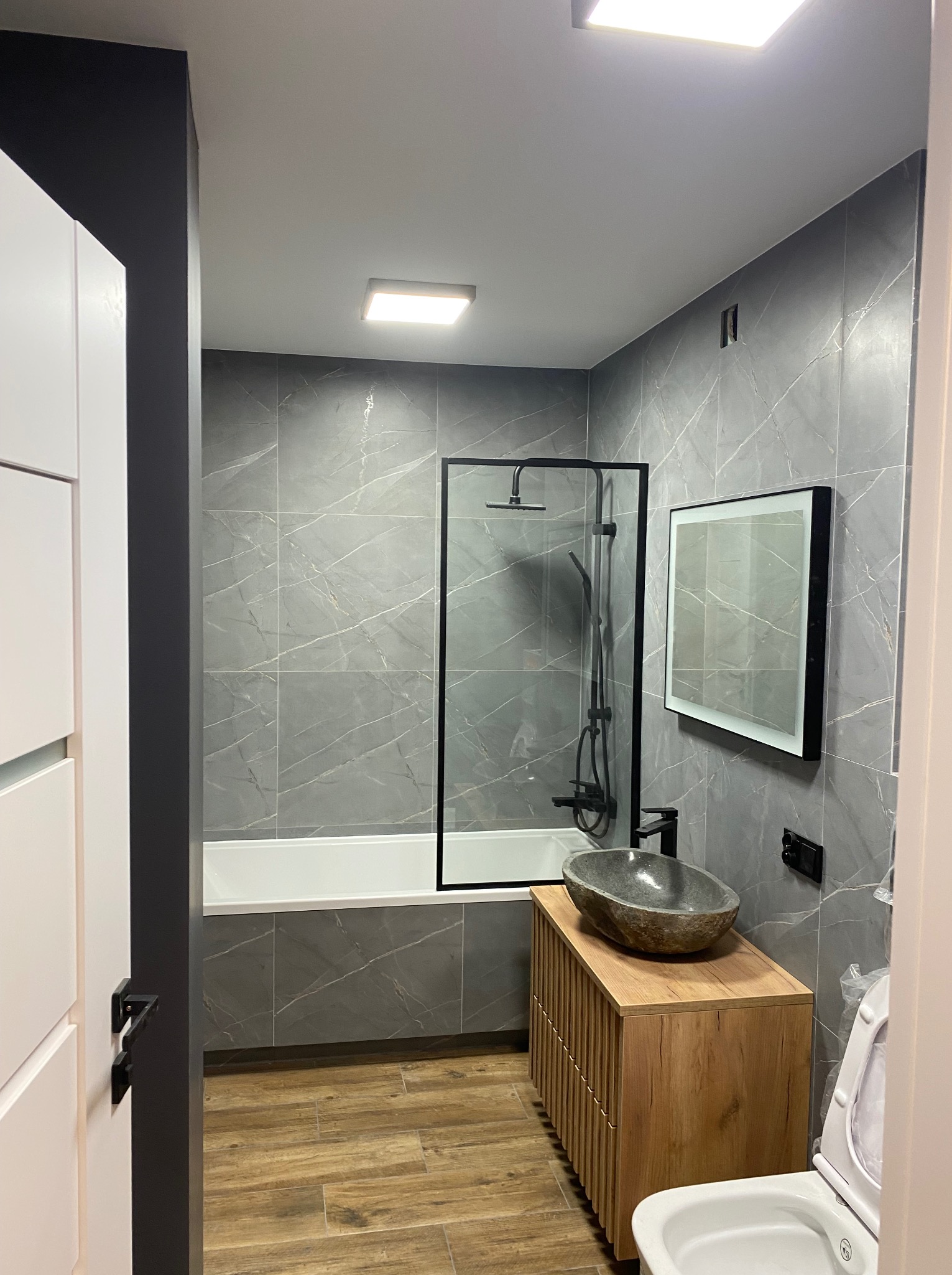Nowoczesna łazienka z szarymi płytkami imitującymi marmur, drewnianą szafką pod umywalką i czarną armaturą. Kabina prysznicowa z czarną ramą.