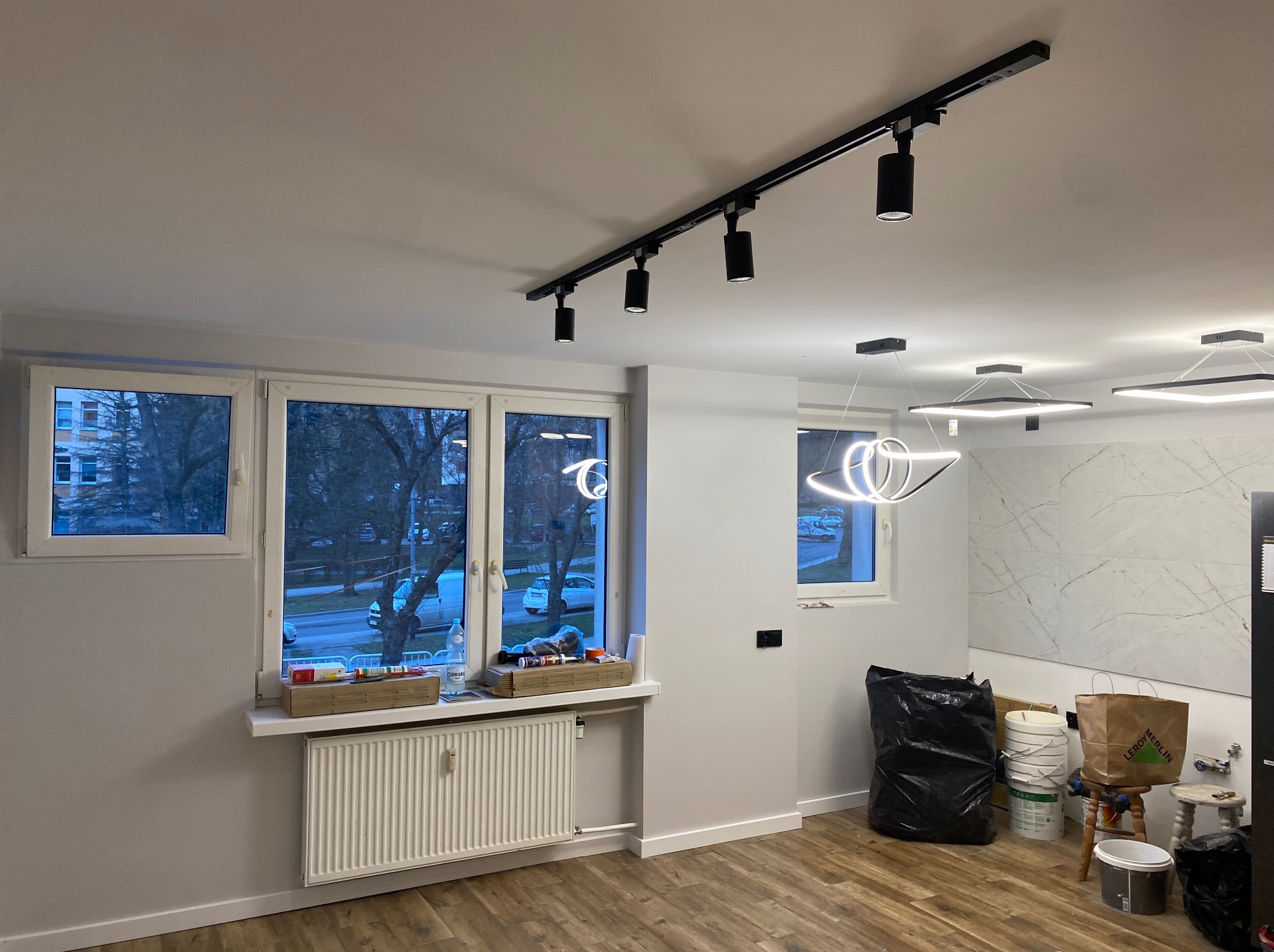 Wykończone wnętrze pokoju z drewnianą podłogą, białymi ścianami, grzejnikiem pod oknem i nowoczesnym oświetleniem sufitowym. Widoczne worki i wiadra po remoncie.