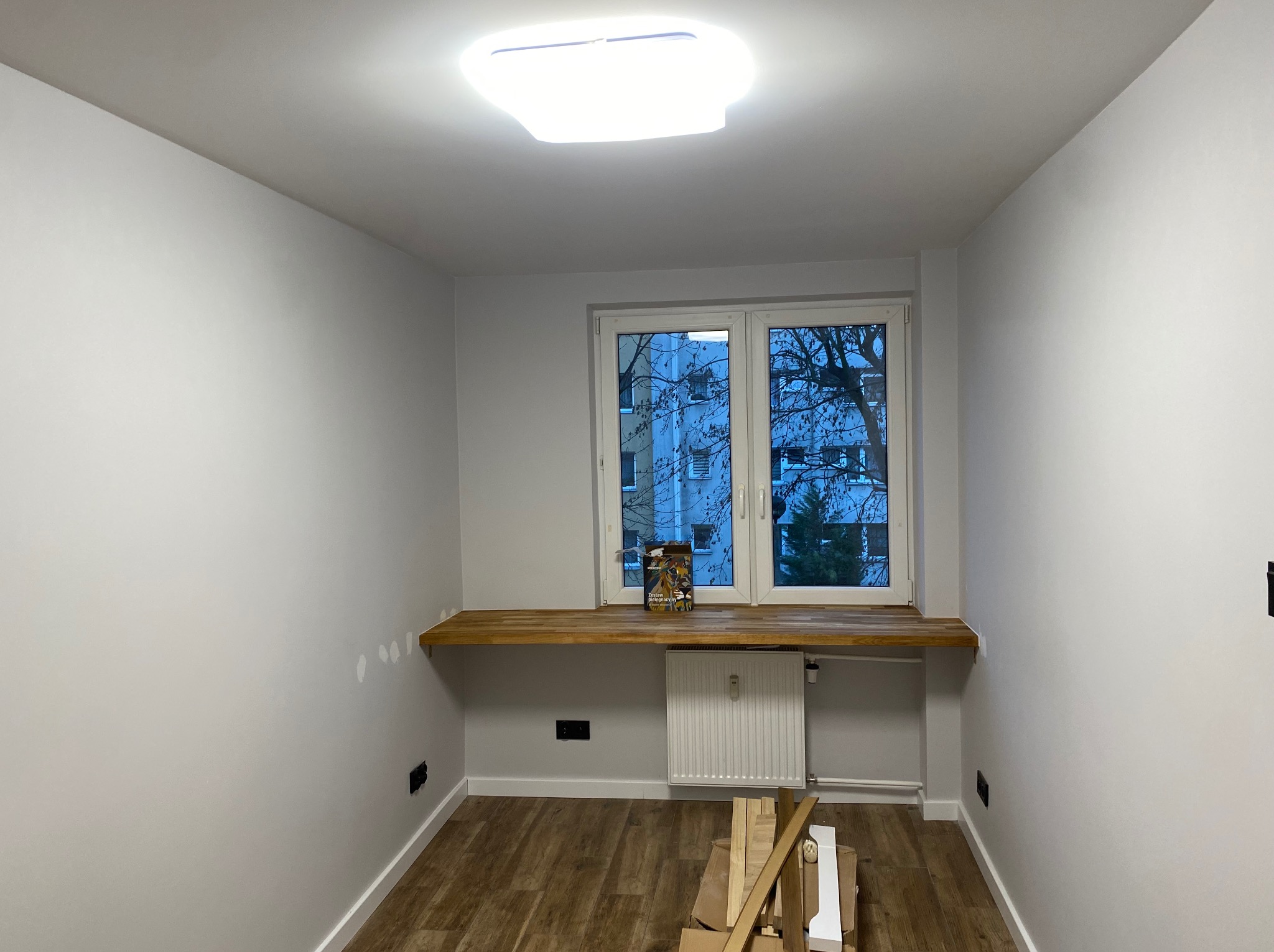 Jasne wnętrze po remoncie: świeżo pomalowane ściany, drewniany blat pod oknem, nowe panele podłogowe. Widoczne materiały budowlane.