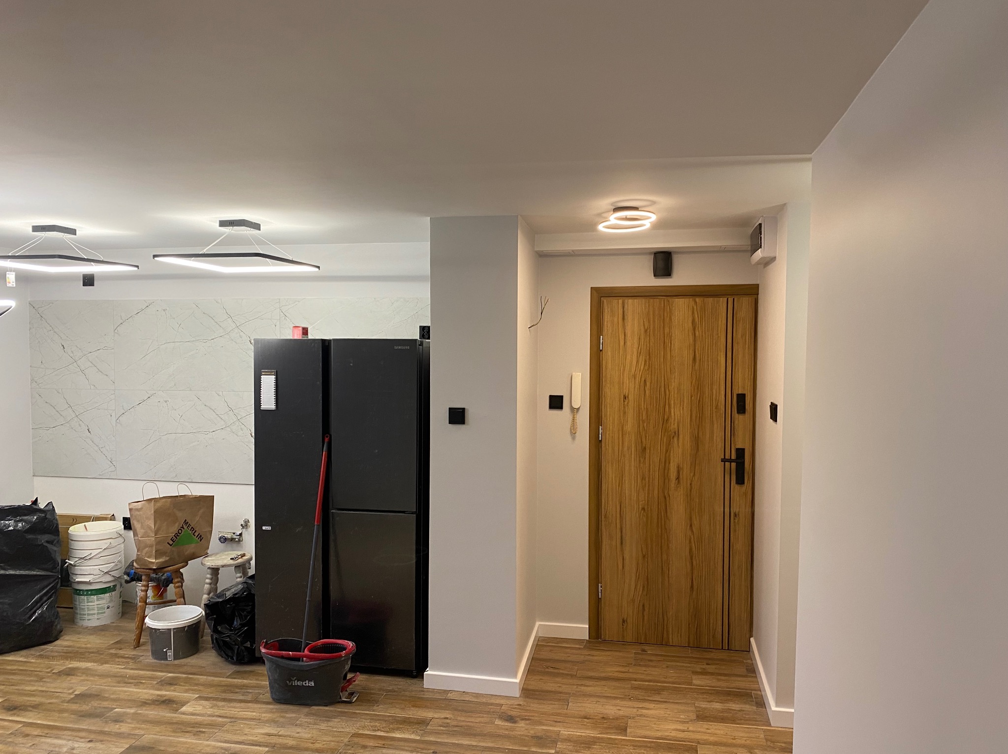 Jasne wnętrze po remoncie: drewnopodobne panele, białe ściany, nowoczesne oświetlenie. Widoczne wiadro i mop sugerują prace porządkowe po zakończeniu prac.