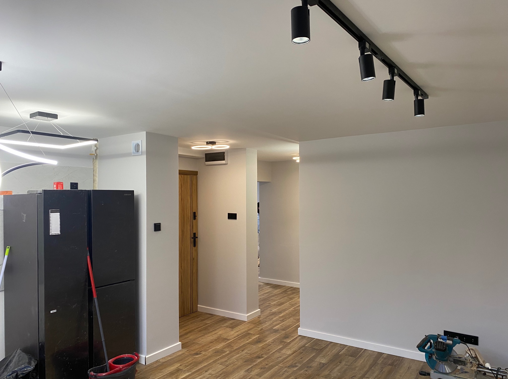 Jasne wnętrze po remoncie: drewnopodobne panele, szare ściany z białymi listwami, nowoczesne oświetlenie sufitowe. W tle widoczne drzwi i kolejne pomieszczenia.