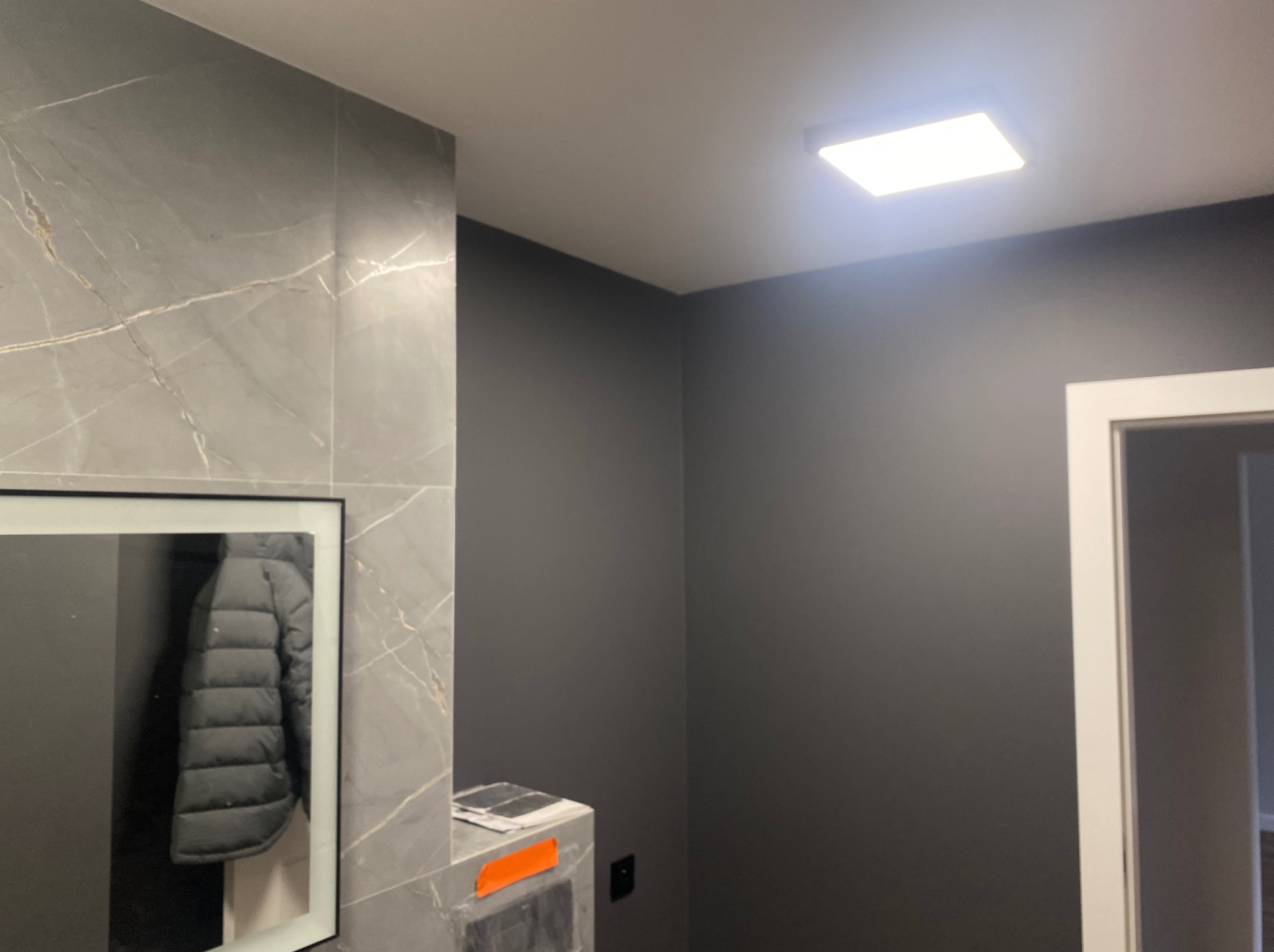 Szara łazienka z marmurowymi płytkami, lustrem odbijającym kurtkę i kwadratową lampą sufitową. Minimalistyczny design, ciemne ściany, białe obramowanie drzwi.