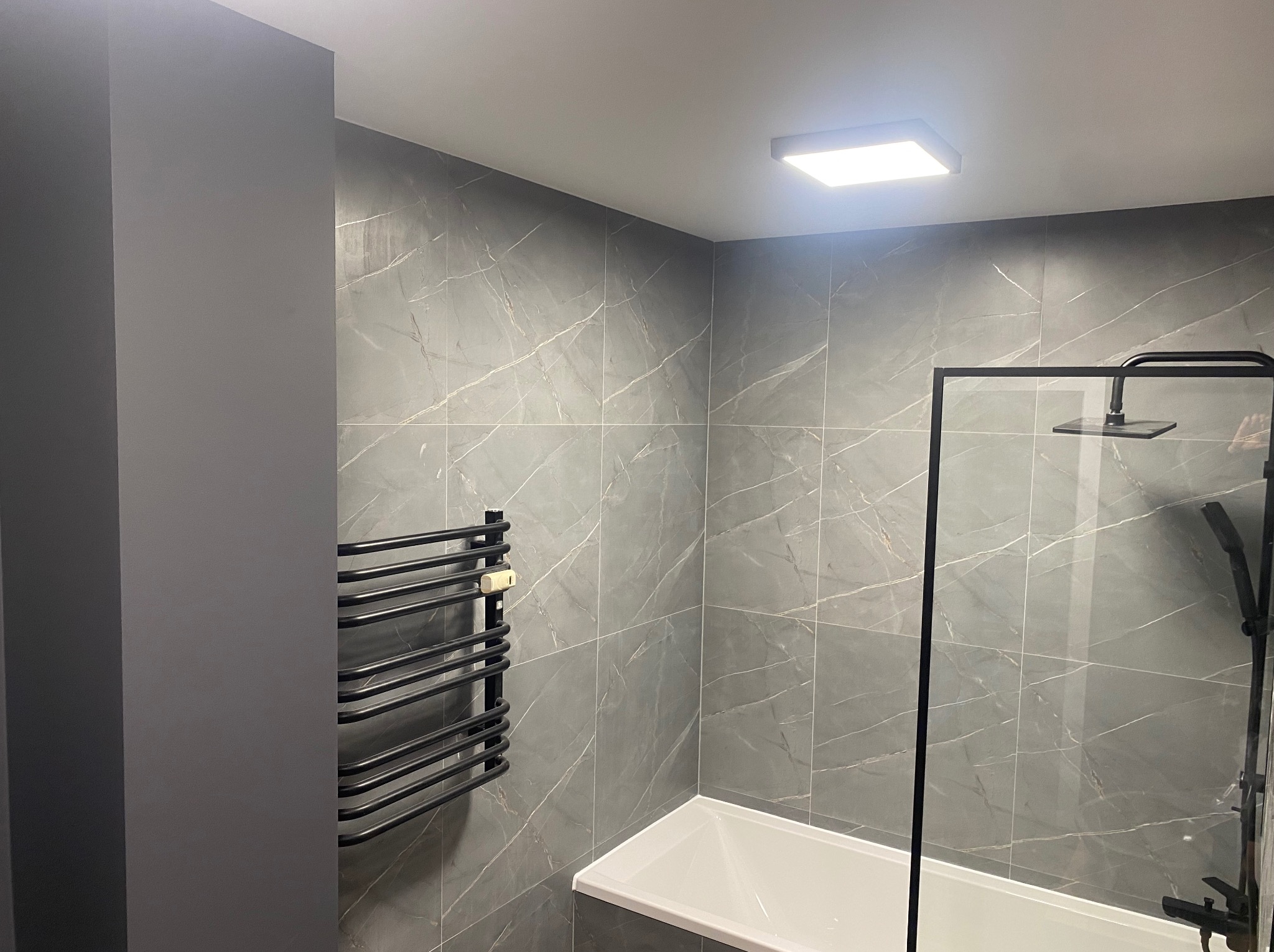 Nowoczesna łazienka z szarymi płytkami imitującymi marmur, czarnym grzejnikiem drabinkowym i kabiną prysznicową typu walk-in z czarną armaturą.