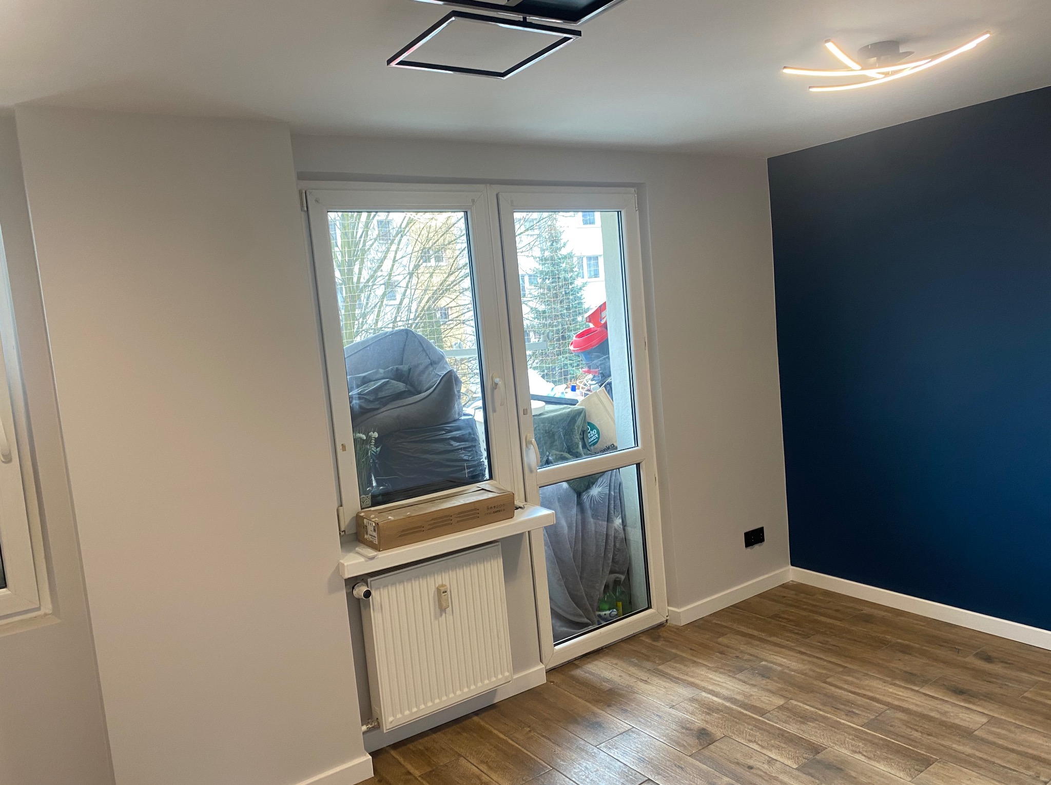 Pokój po odświeżeniu: drewnopodobne panele, szara ściana i granatowa ściana akcentująca. Okno z widokiem na zewnątrz, grzejnik pod oknem. Nowoczesne oświetlenie sufitowe.