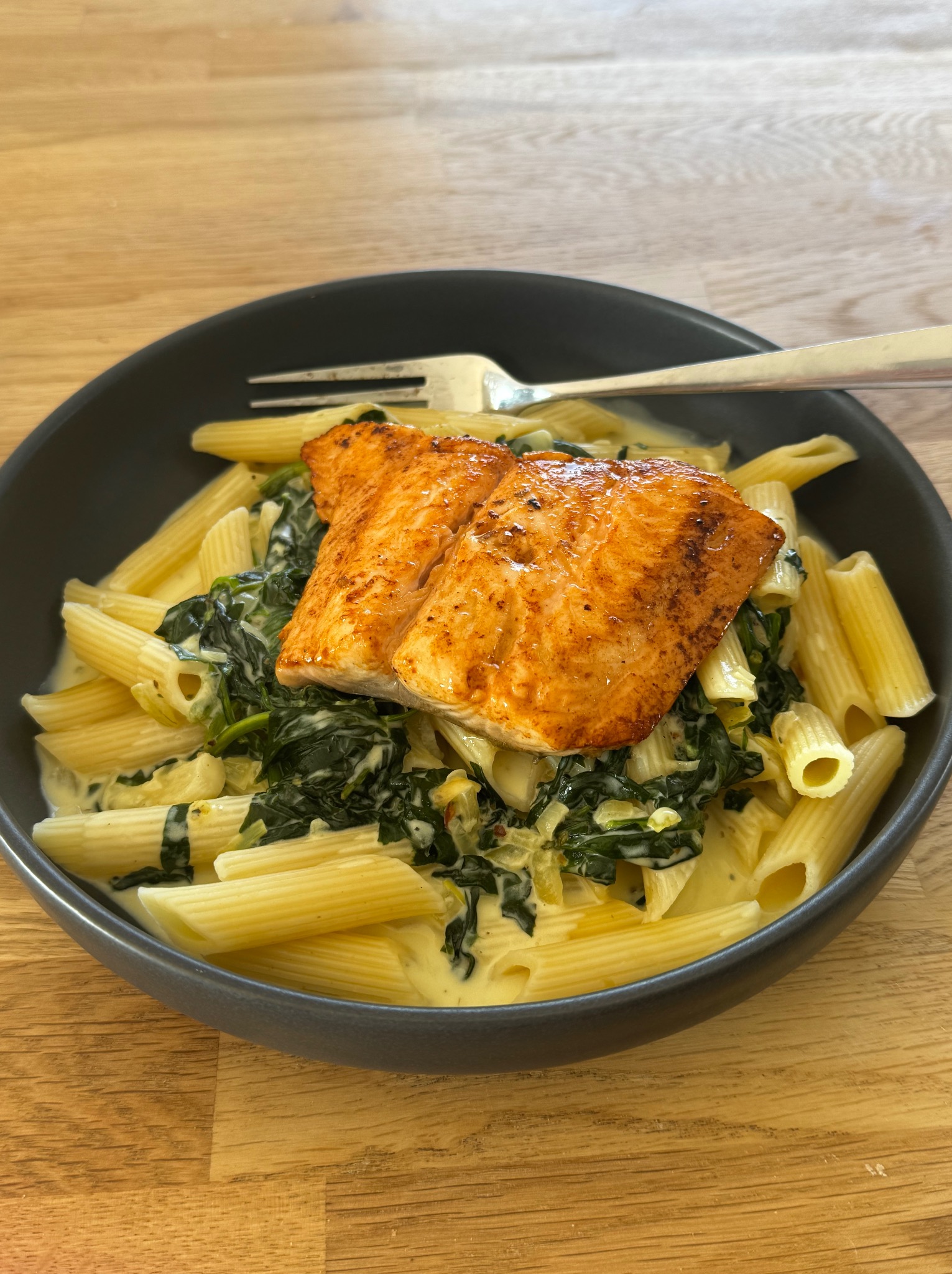 Penne z łososiem i szpinakiem w kremowym sosie, podane w ciemnej misce z widelcem na drewnianym stole. Danie gotowe do spożycia, oświetlone naturalnym światłem.