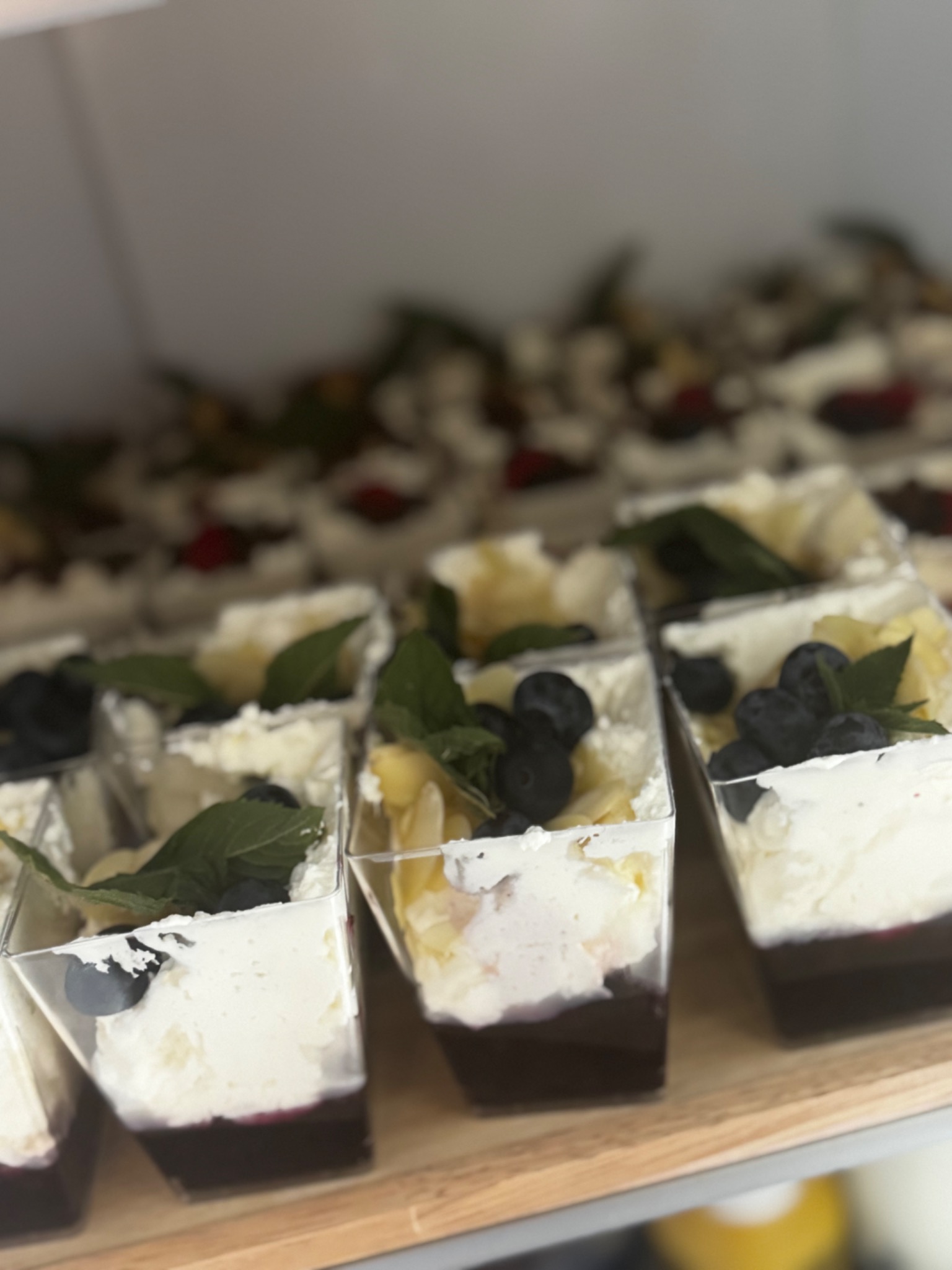 Deser w przezroczystych pojemnikach: krem, owoce (borówki, ananas), listki mięty. Ułożone rzędami na drewnianej tacy. Widok z góry, lekki kąt.