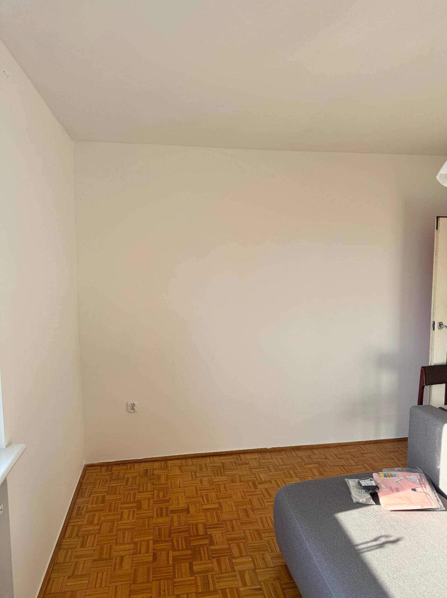 Pusty pokój z parkietem w jodełkę, świeżo pomalowanymi ścianami na biało i szarą sofą w rogu. Widoczny kontakt elektryczny i fragment drzwi.