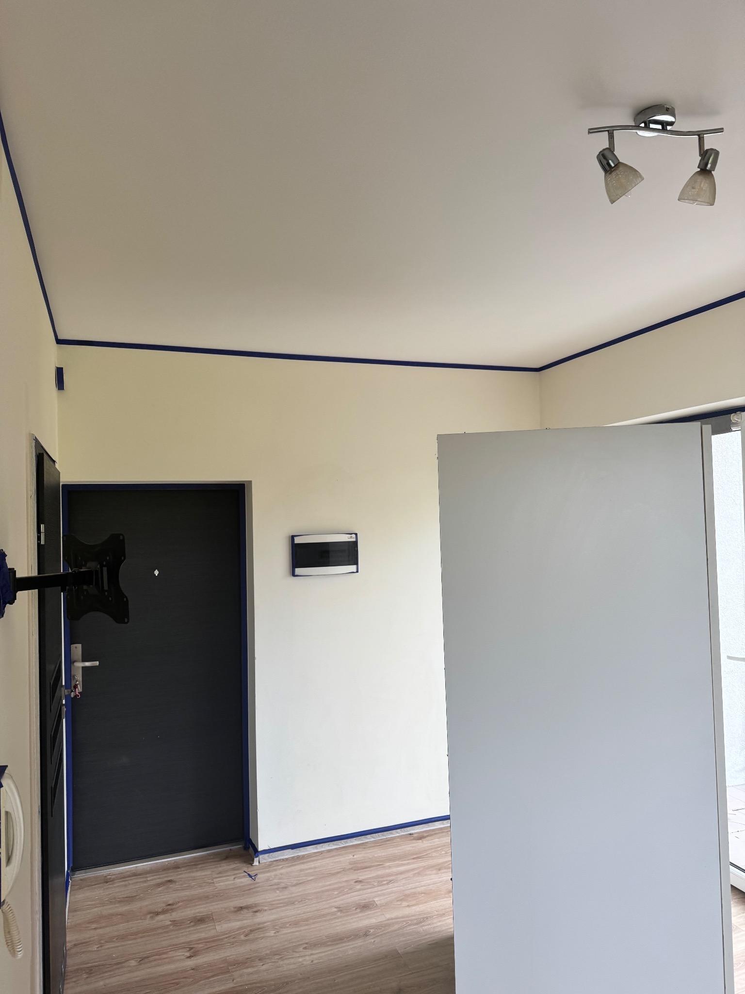 Przygotowane do malowania wnętrze: ściany i sufit zabezpieczone niebieską taśmą malarską, widoczna lampa sufitowa i drzwi wejściowe.