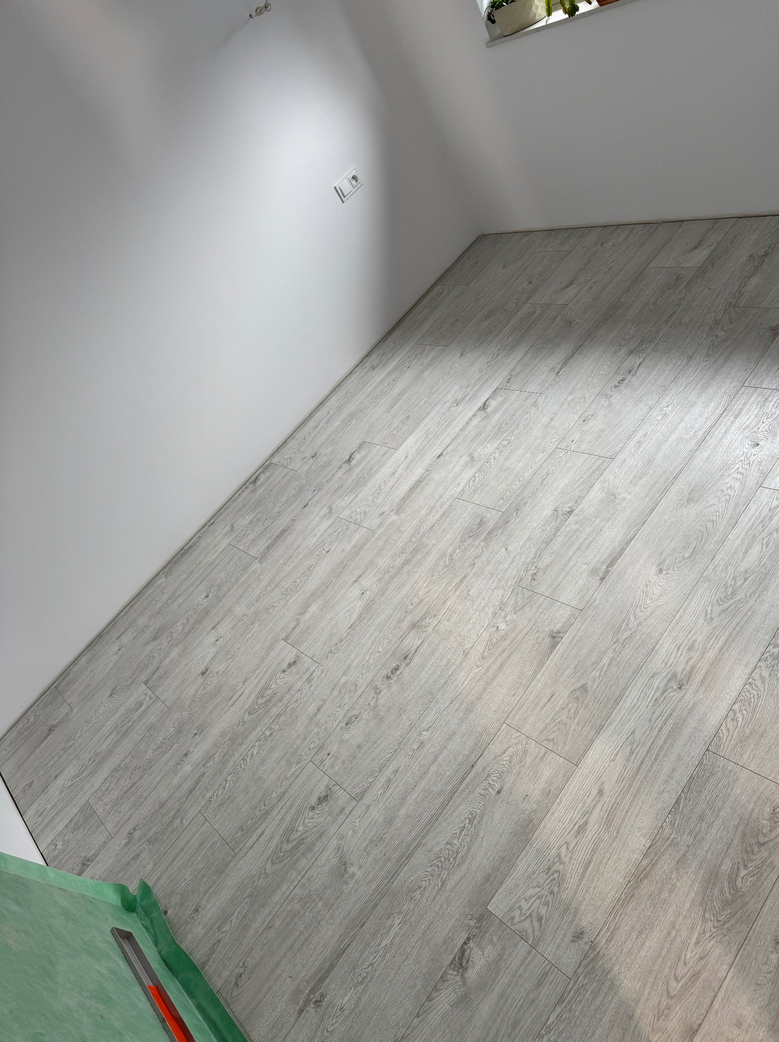 Jasne panele podłogowe ułożone w rogu pokoju, widoczne narzędzia montażowe i fragment ściany z gniazdkiem. Minimalistyczne wnętrze w trakcie wykańczania.