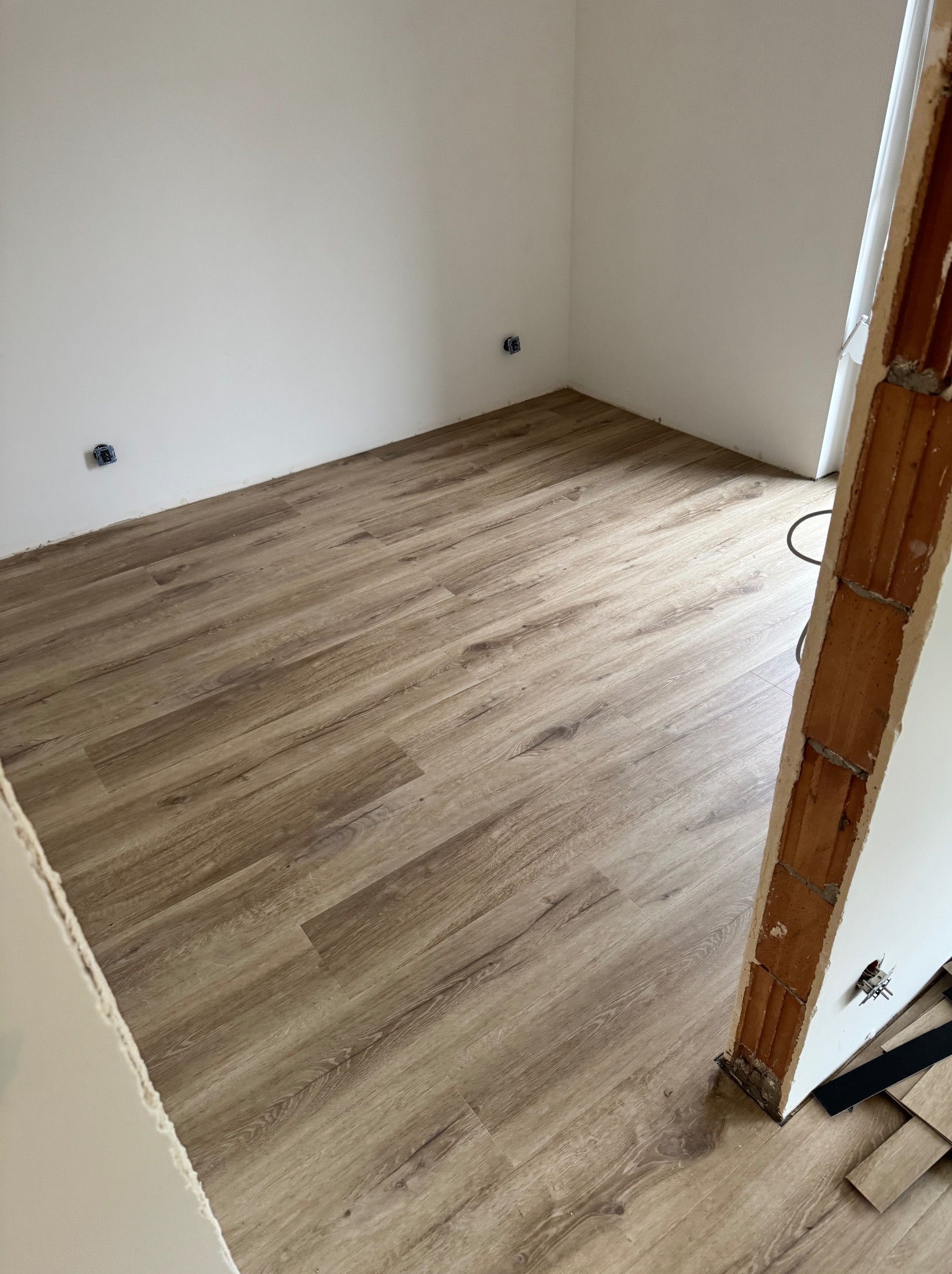 Jasne panele podłogowe ułożone w rogu pokoju z białymi ścianami i widocznymi gniazdkami elektrycznymi. Fragment ościeżnicy z cegły w prawym dolnym rogu.