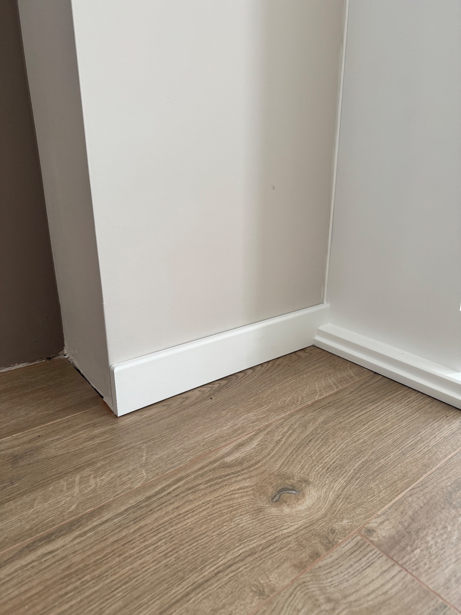 Jasne panele podłogowe z drewnianym wzorem, białe listwy przypodłogowe przy ścianie pomalowanej na jasny odcień. Widoczny fragment narożnika.