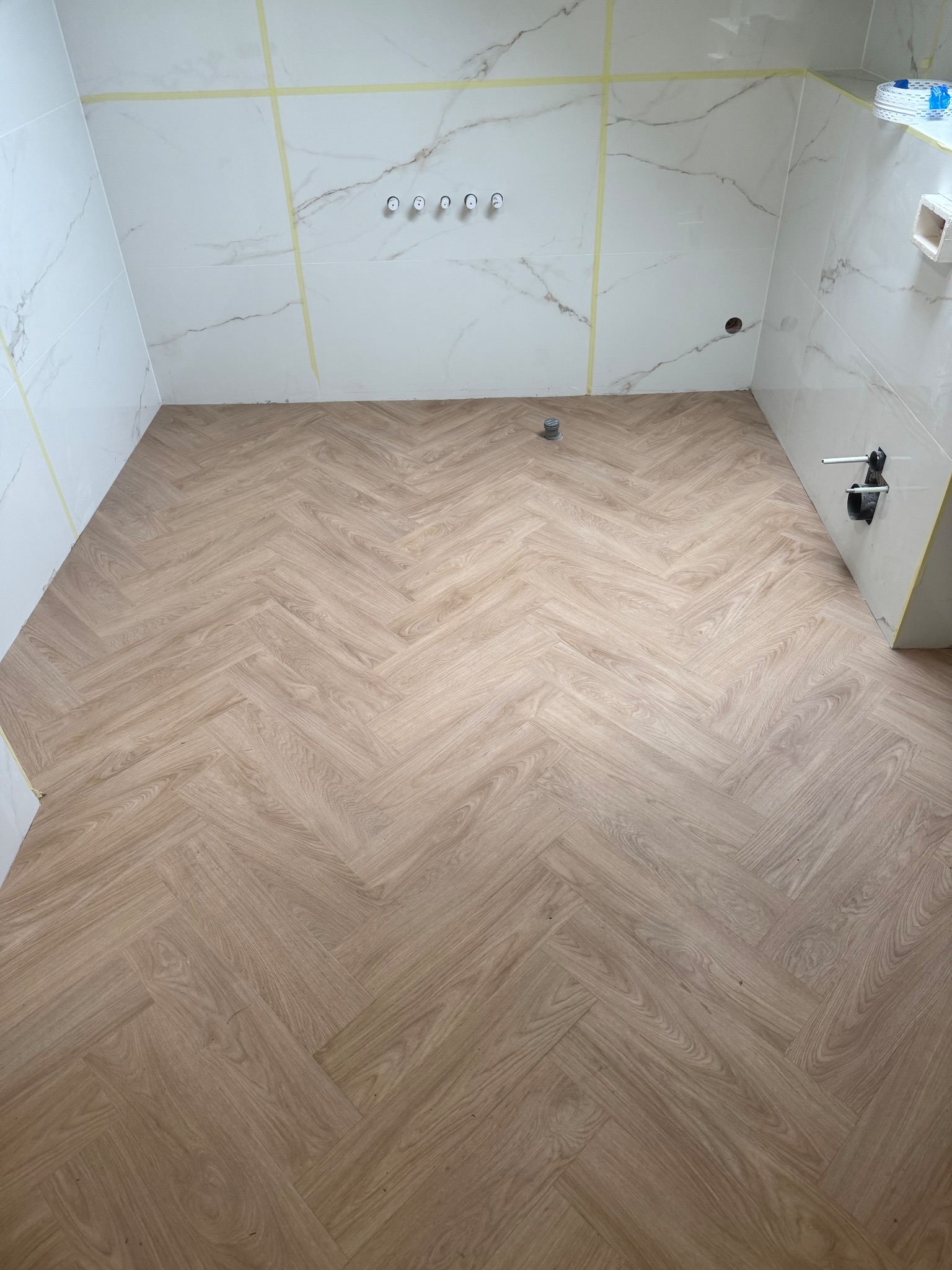 Ułożona jodełka z paneli podłogowych w jasnym odcieniu drewna, w trakcie wykańczania łazienki z białymi płytkami imitującymi marmur i taśmą malarską na ścianach.