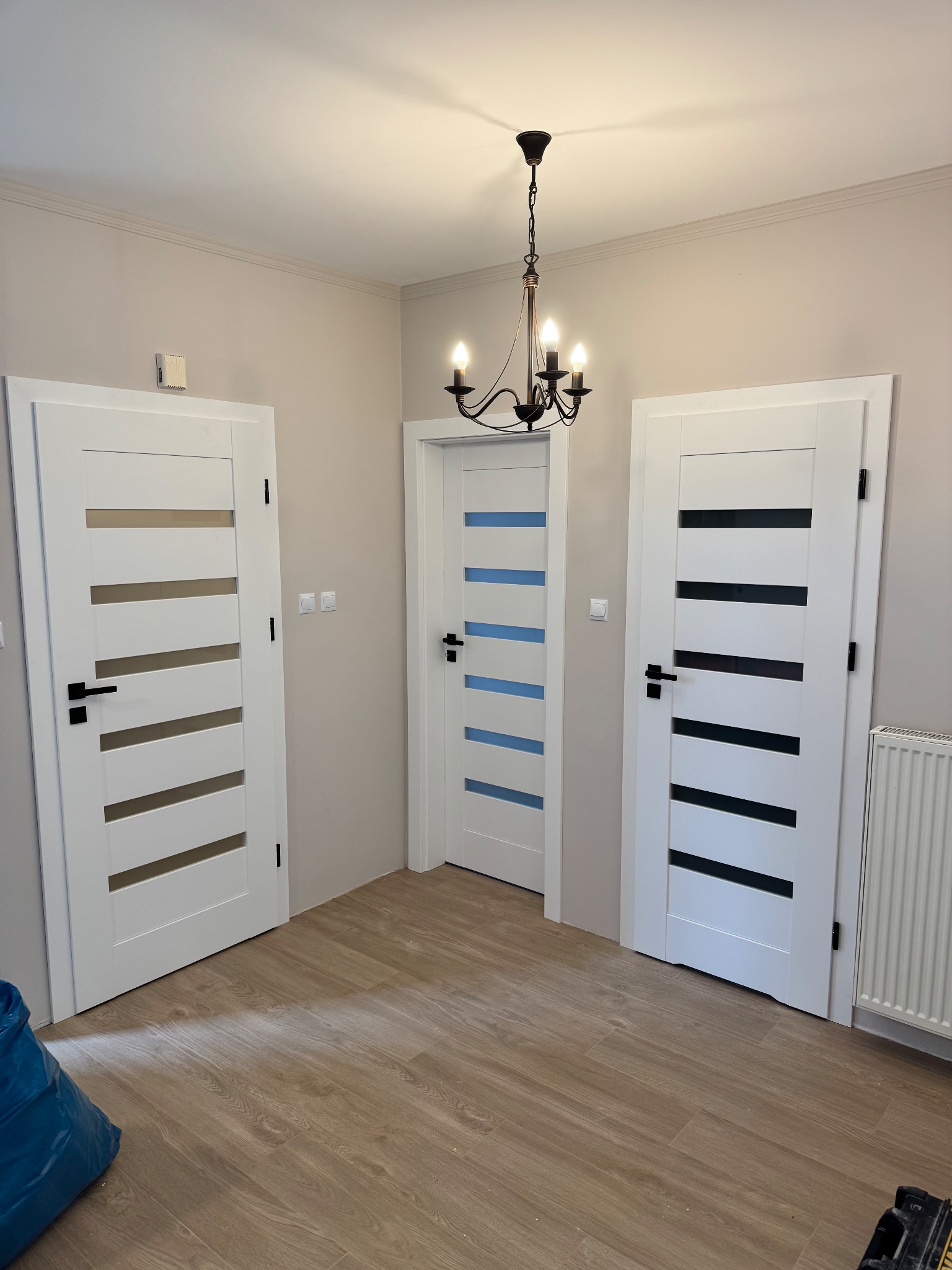 Trzy białe drzwi z przeszkleniami w różnych kolorach, zamontowane w pokoju w Śremie. Widoczna podłoga z paneli i wisząca lampa.