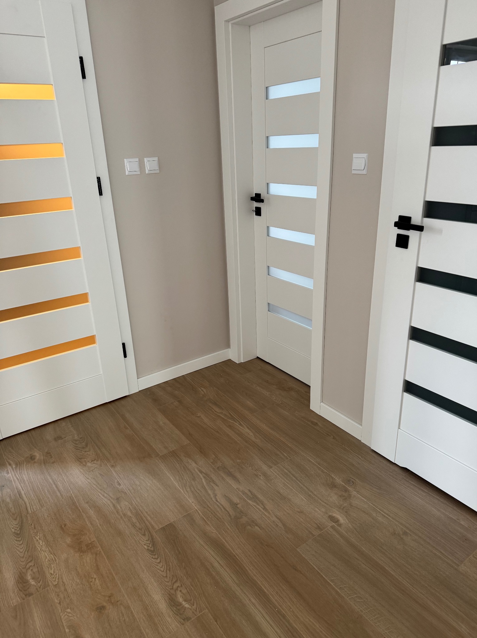 Trzy białe drzwi wewnętrzne z czarnymi klamkami i różnymi wzorami przeszkleń, widoczne na tle beżowej ściany i podłogi z paneli imitujących drewno.