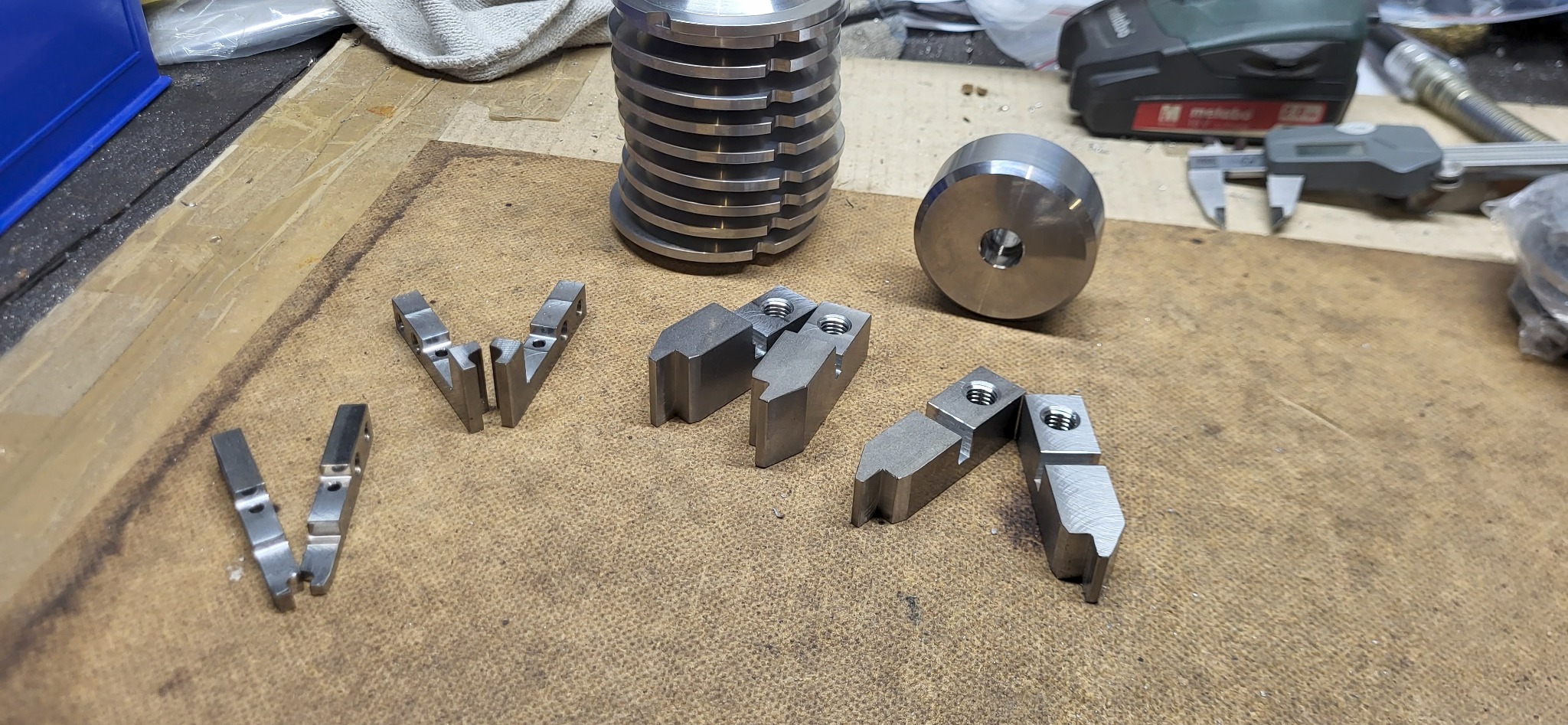 Precyzyjnie wykonane, metalowe elementy tokarskie, w tym uchwyty szczękowe, leżące na warsztatowym stole. Widoczne narzędzia pomiarowe i akumulatorowa wiertarka.