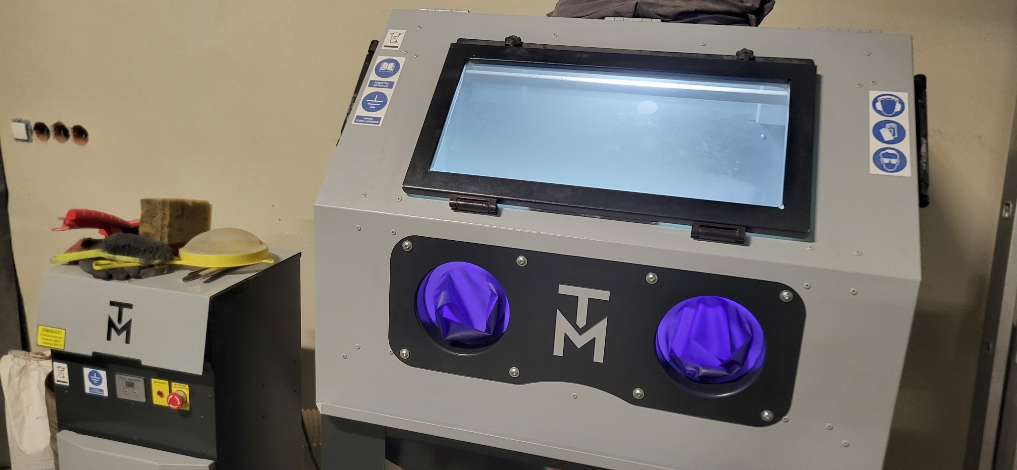 Szara kabina do piaskowania z logo TM, widoczne rękawice ochronne i okno inspekcyjne. Obok stoi mniejsza maszyna z przyciskami i narzędziami na górze.