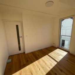 Ruslan Reshko Reshko Bud - Jasne, wykończone wnętrze pokoju z drewnianą podłogą, białymi ścianami, drzwiami z przeszkleniem i oknem z widokiem na budynek. Promienie słoneczne na podłodze.