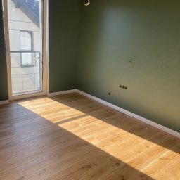 Ruslan Reshko Reshko Bud - Pomalowane na oliwkowo wnętrze pokoju z drewnianą podłogą, listwami przypodłogowymi i oknem balkonowym. Widoczne gniazdka elektryczne i ślady po instalacji karnisza.