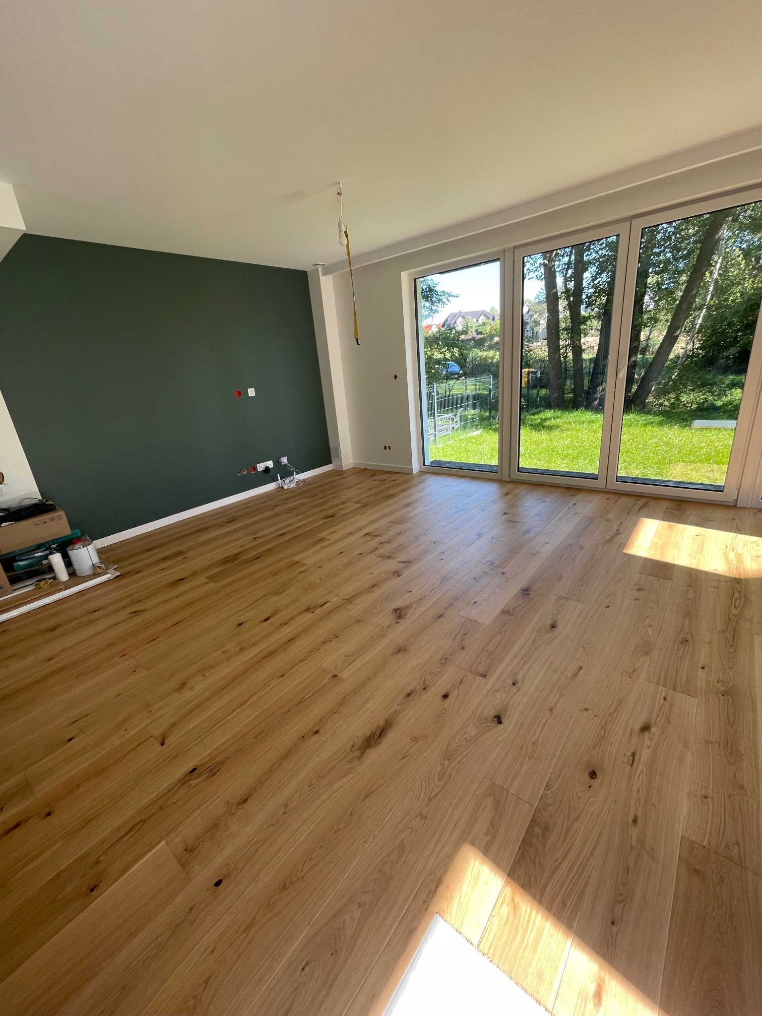 Jasna drewniana podłoga w nowym pokoju z dużymi oknami i ciemnozieloną ścianą. Widoczne gniazdka elektryczne i listwy przypodłogowe. Częściowo widoczne narzędzia.