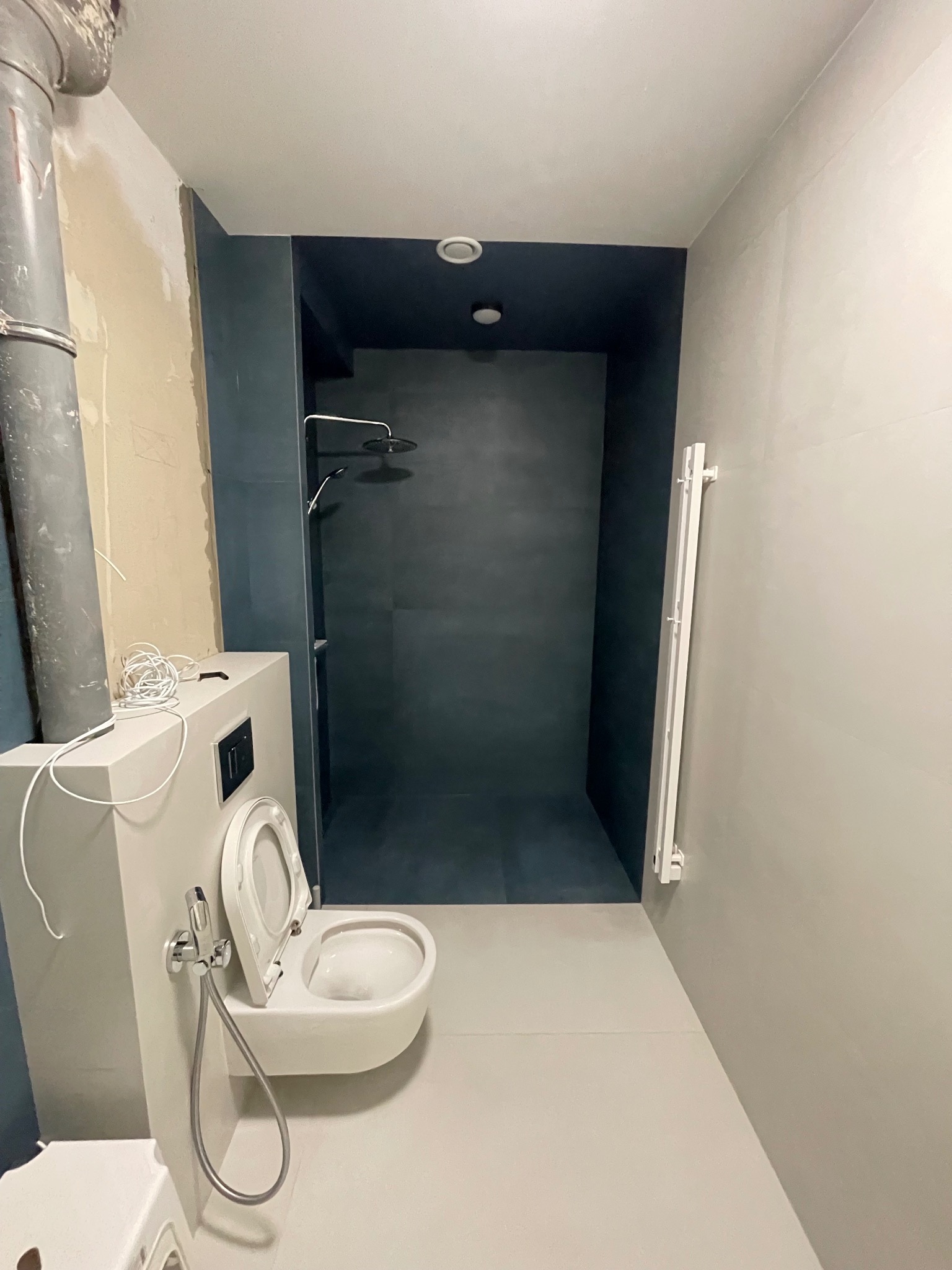 Nowoczesna łazienka z kabiną typu walk-in w odcieniach szarości i granatu. Widoczna podtynkowa toaleta z bidetką i grzejnik drabinkowy. Częściowo wykończone ściany.