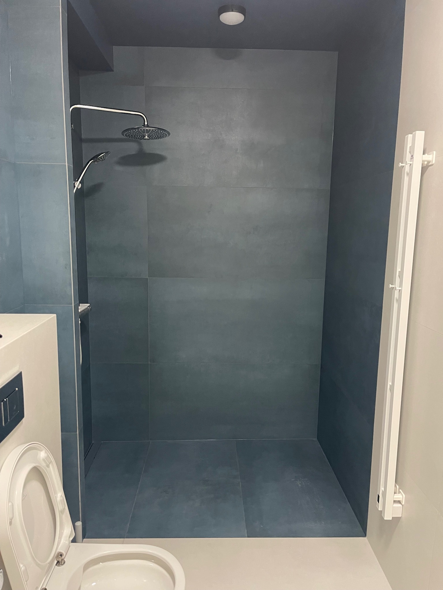 Nowoczesna kabina prysznicowa z deszczownicą i odpływem liniowym, wykończona płytkami w odcieniach szarości. Widoczny fragment toalety i grzejnik drabinkowy.