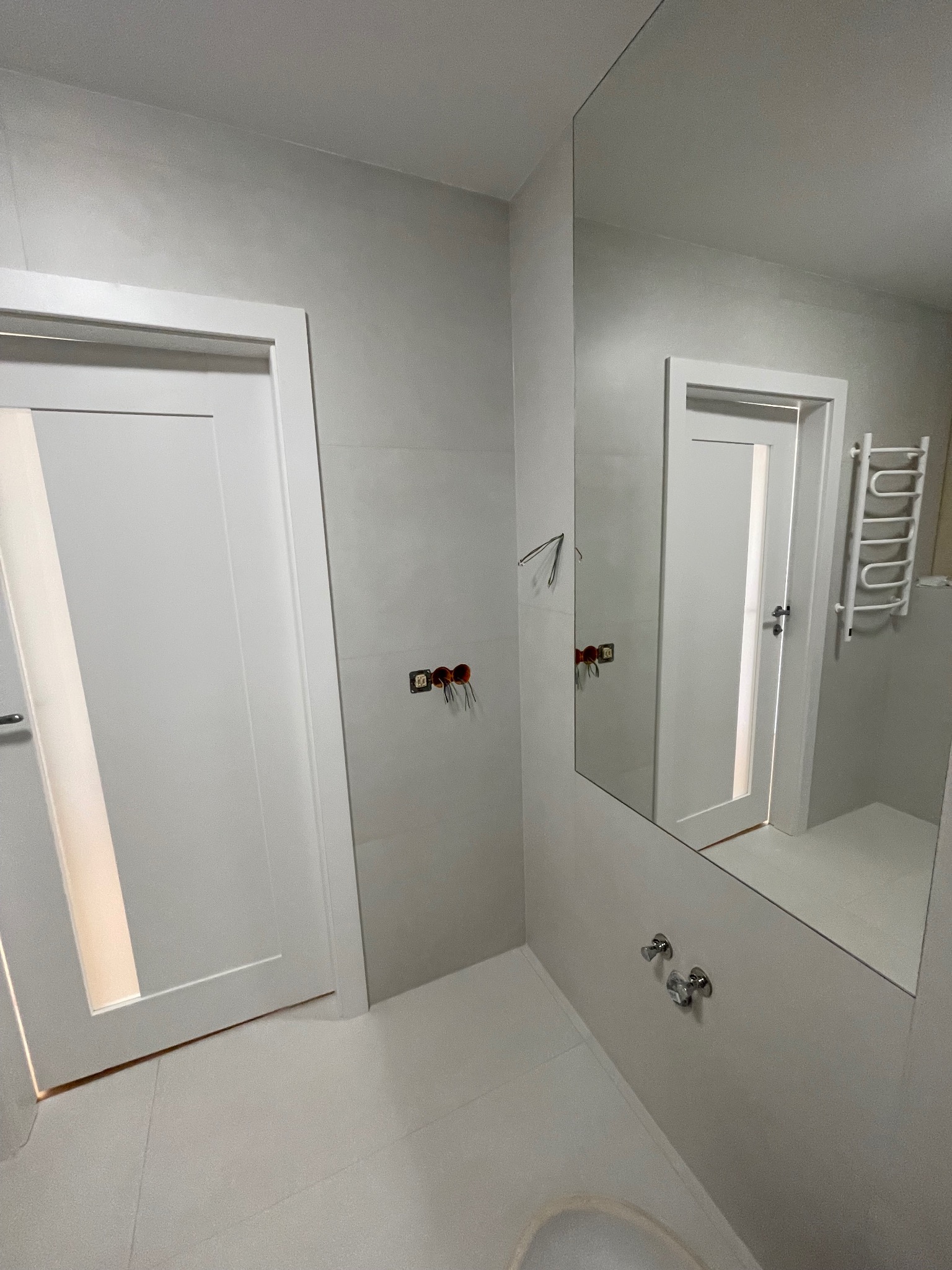 Wykończona łazienka w odcieniach szarości z białymi drzwiami i grzejnikiem drabinkowym, widoczna w lustrze. Minimalistyczny design, płytki na ścianach i podłodze.