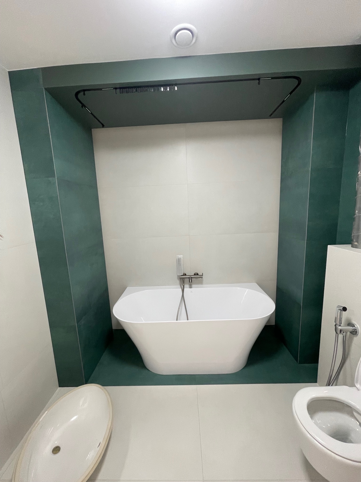 Nowoczesna łazienka z białą wanną wolnostojącą, zielonymi akcentami i minimalistycznym oświetleniem. Uwagę zwraca nietypowe wykończenie wnęki w odcieniach zieleni.