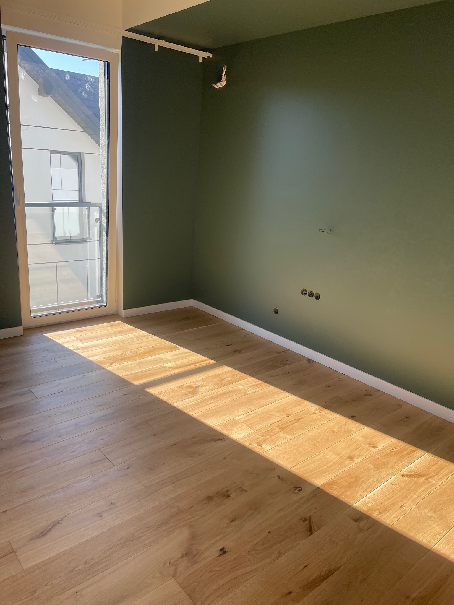 Pomalowane na oliwkowo wnętrze pokoju z drewnianą podłogą, listwami przypodłogowymi i oknem balkonowym. Widoczne gniazdka elektryczne i ślady po instalacji karnisza.