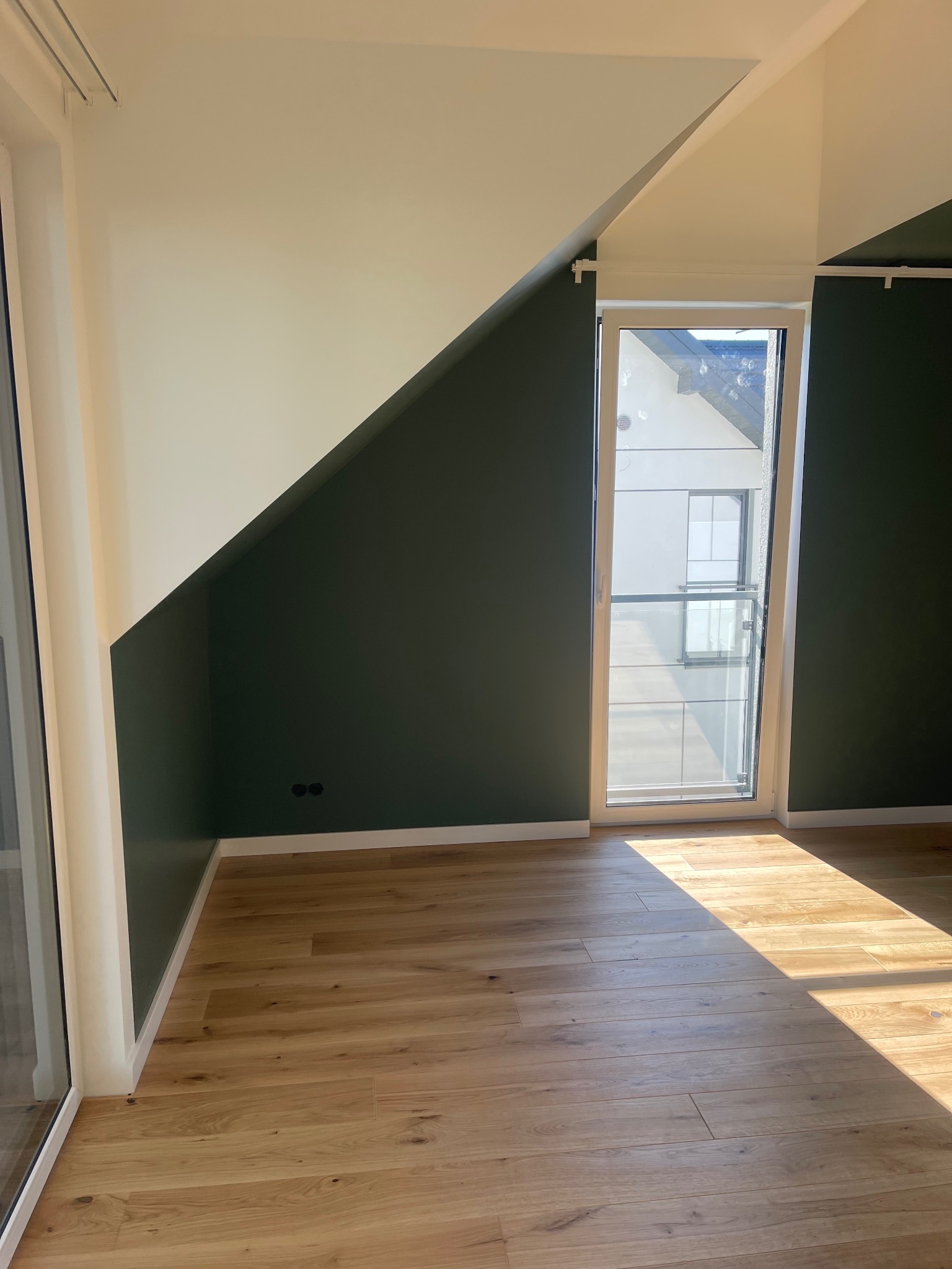 Pomieszczenie po malowaniu: ściany w kolorze butelkowej zieleni i białe, drewniana podłoga, okno balkonowe. Widoczne gniazdka elektryczne.