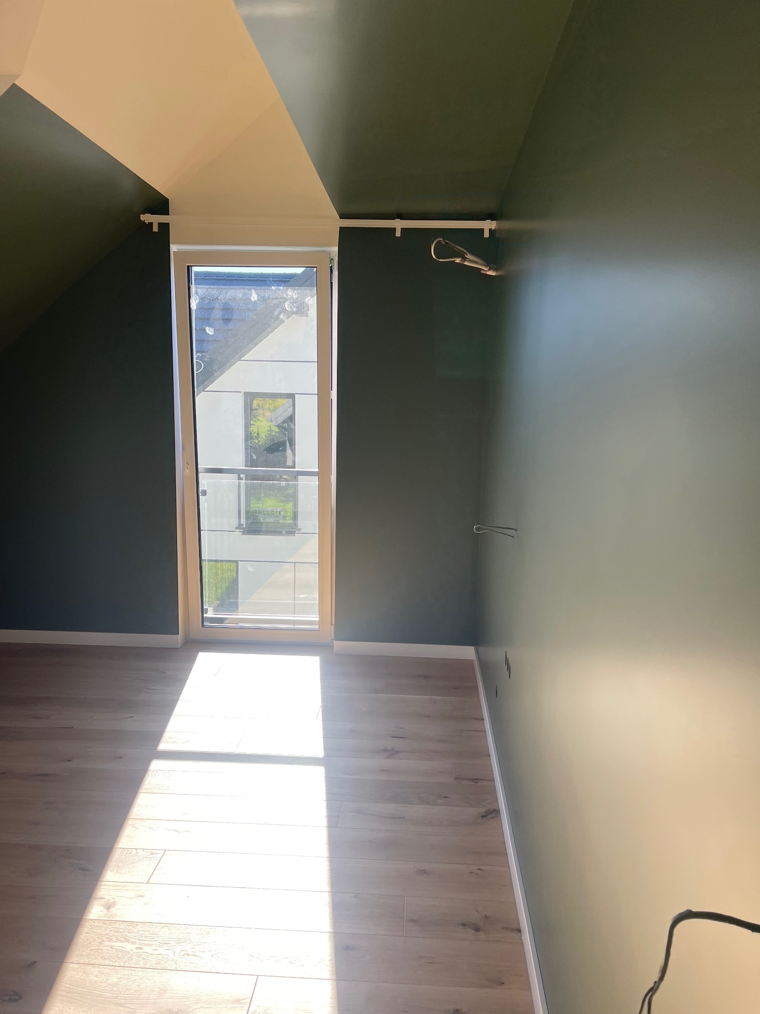 Pomieszczenie na poddaszu po malowaniu ścian na ciemnozielony kolor. Widoczne okno z białymi ramami i drewniana podłoga, na którą pada snop światła.