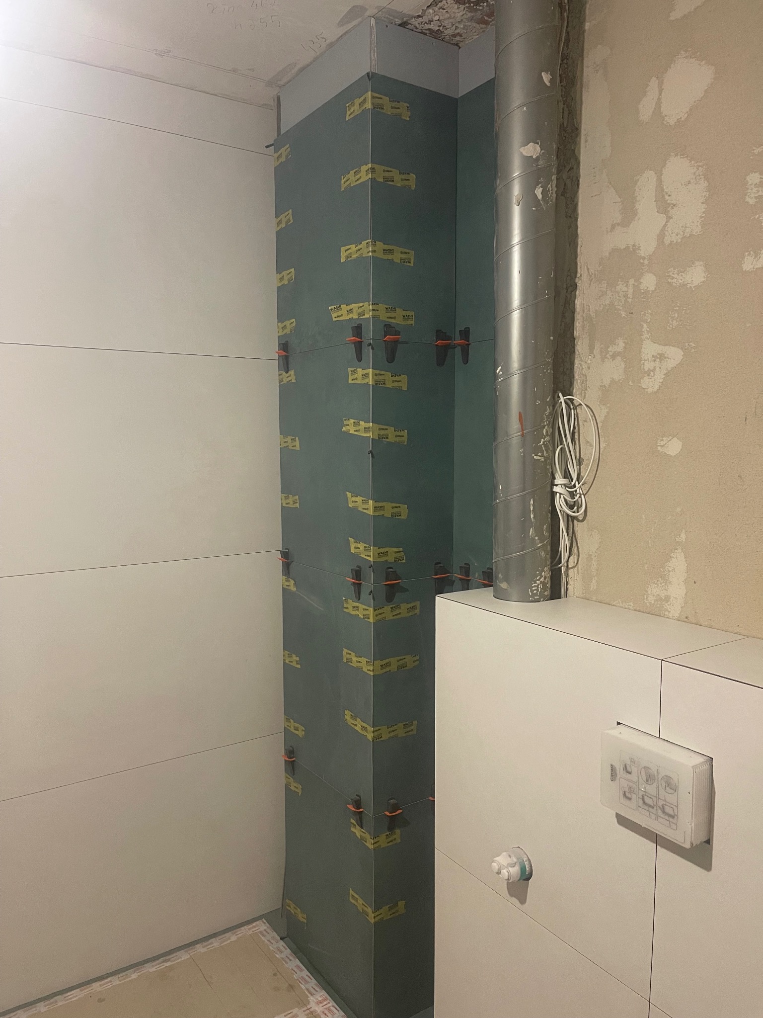 Układanie płytek na ścianie w łazience w Markach. Widoczne płytki, pion kanalizacyjny i system poziomowania płytek. Prace w toku, surowy stan.