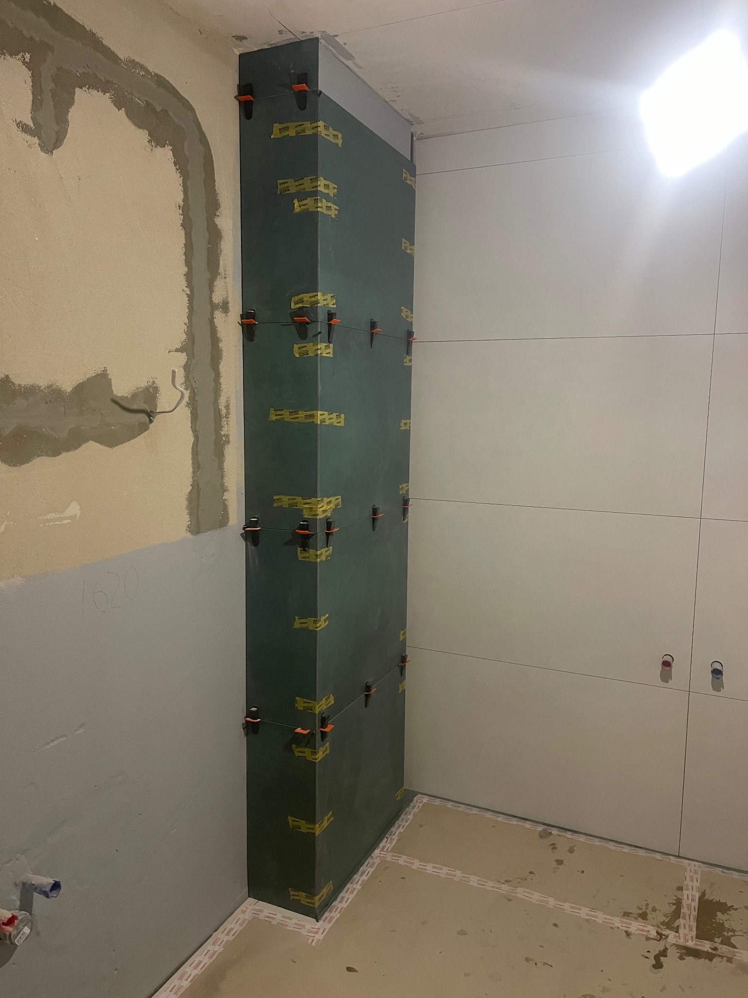 Wyrównywanie narożnika płytami budowlanymi z użyciem klipsów i taśmy. Ściana z jasnymi płytkami w tle. Widoczne ślady po szpachlowaniu na ścianie obok.