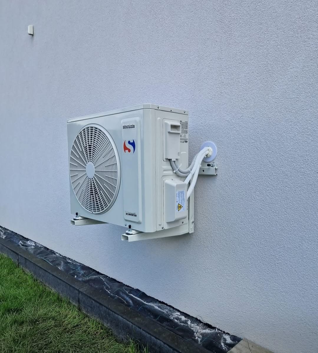 Biała jednostka zewnętrzna klimatyzacji Sinclair zamontowana na elewacji budynku, widoczne przewody i elementy montażowe. Ujęcie z dołu, trawa w tle.