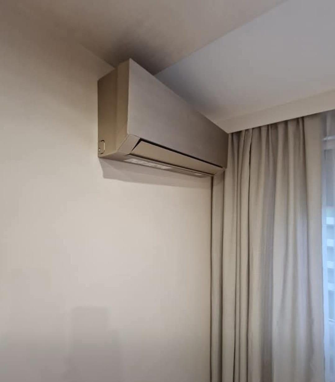 Beżowy klimatyzator ścienny w jasnym wnętrzu, widoczny fragment zasłony. Minimalistyczny design urządzenia, montaż przy suficie.