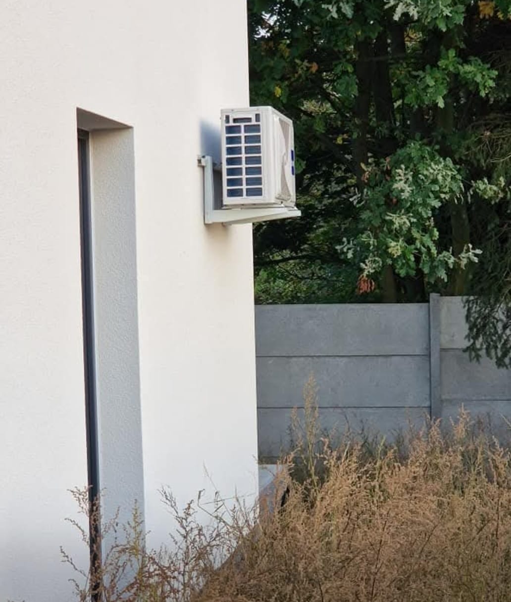 Biała jednostka zewnętrzna klimatyzacji zamontowana na ścianie budynku z widocznym fragmentem okna i zieleni w tle. Ujęcie z dołu, podkreślające montaż.