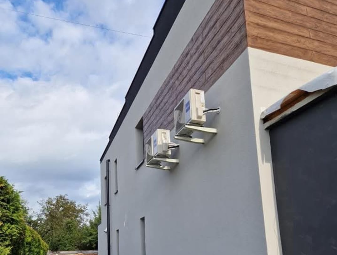 Elewacja budynku z trzema zamontowanymi klimatyzatorami, widoczne przewody. Część budynku wykończona panelami imitującymi drewno. Błękitne niebo w tle.