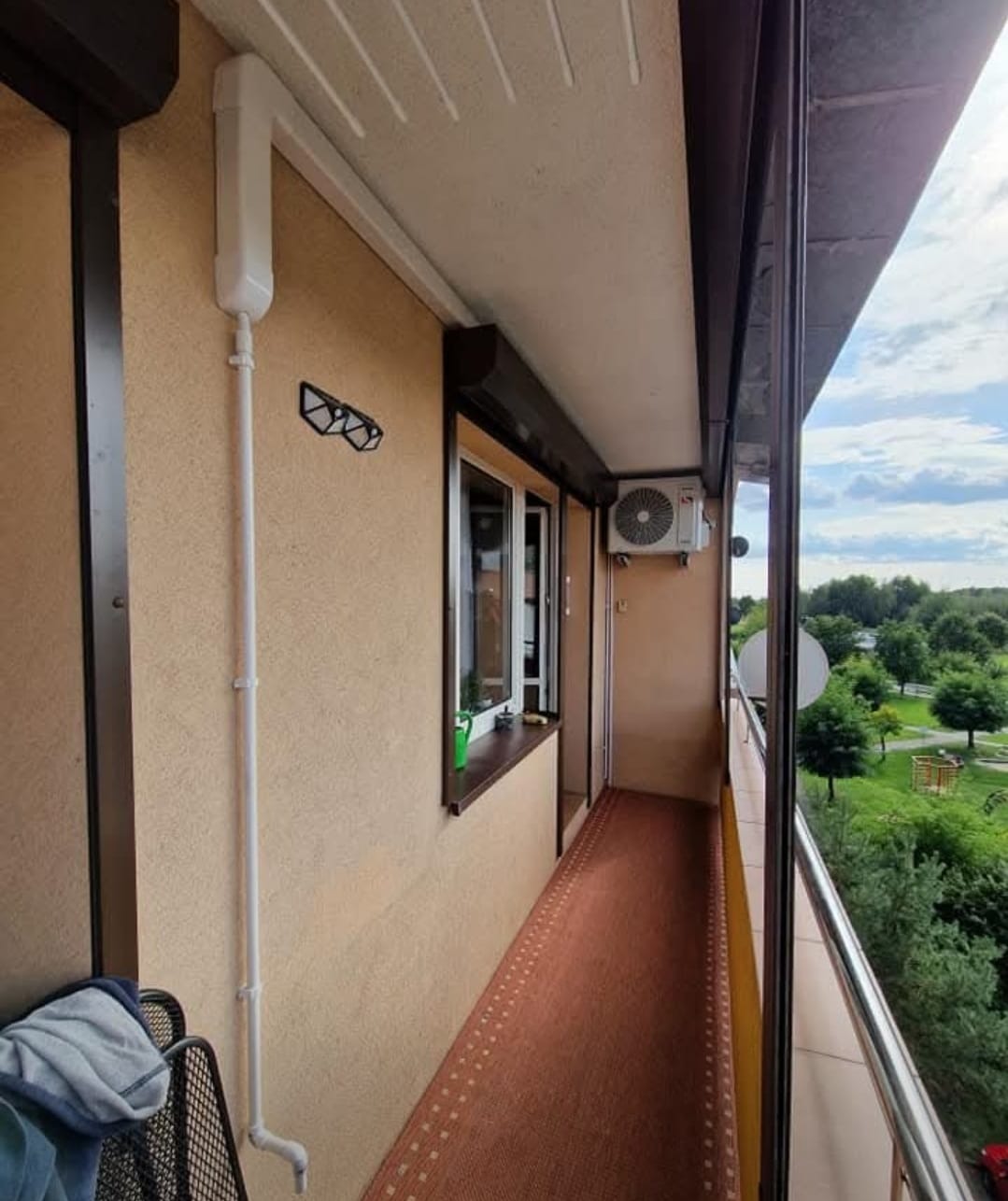 Kompaktowa klimatyzacja na balkonie z brązową ramą okienną i widokiem na zieleń. Biała rura odprowadzająca skropliny. Dywanik w odcieniach brązu.