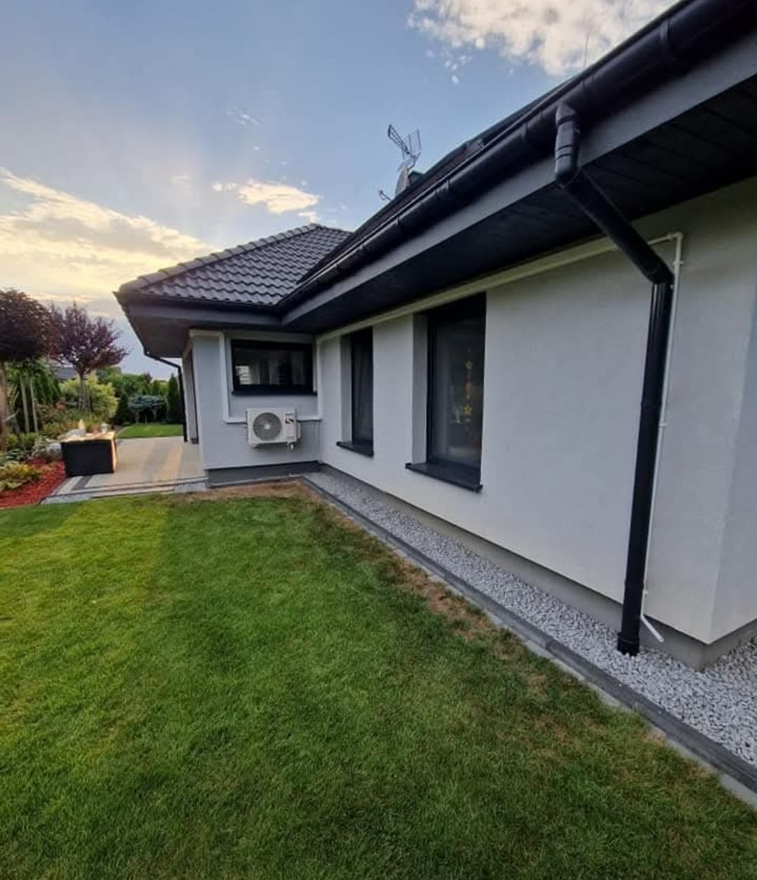 Biały dom z zamontowaną klimatyzacją na ścianie, widok z zewnątrz. Czarny system rynnowy i okna. Zadbane otoczenie z trawnikiem i elementami dekoracyjnymi.