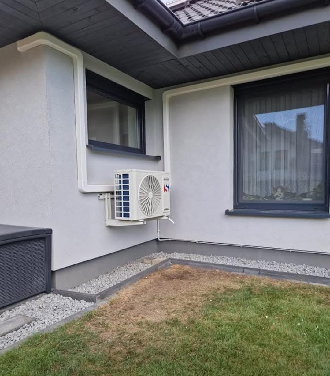 Biała jednostka zewnętrzna klimatyzacji zamontowana na elewacji domu, z widocznymi rurami instalacyjnymi. Obok okno z ciemną ramą i fragment trawnika.