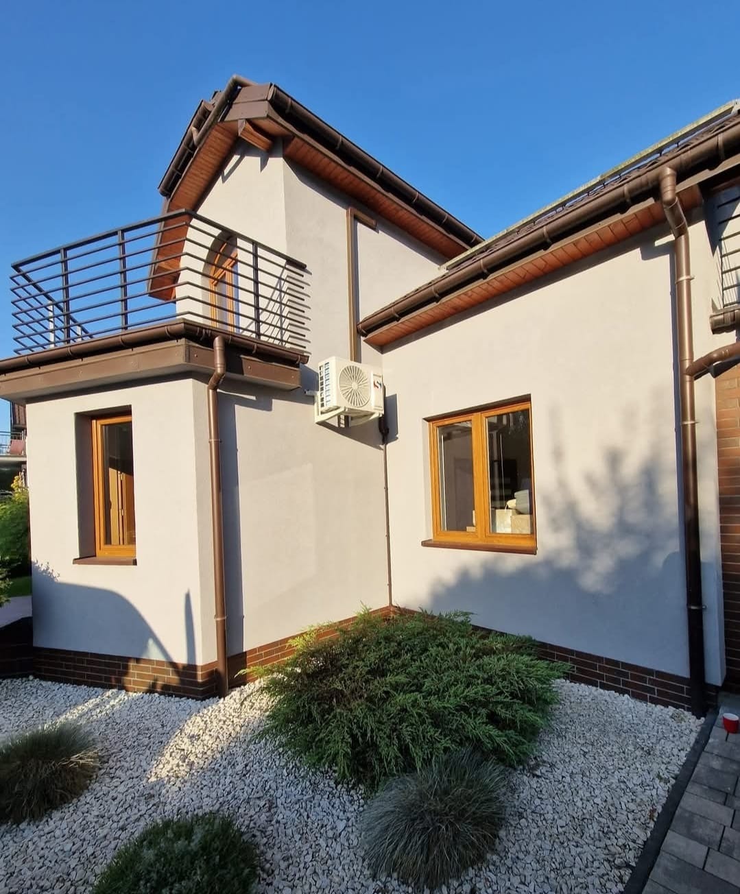Biała elewacja domu z brązowymi detalami, widoczna jednostka zewnętrzna klimatyzacji, balkon z metalową balustradą i ozdobny ogródek z białym kamieniem.