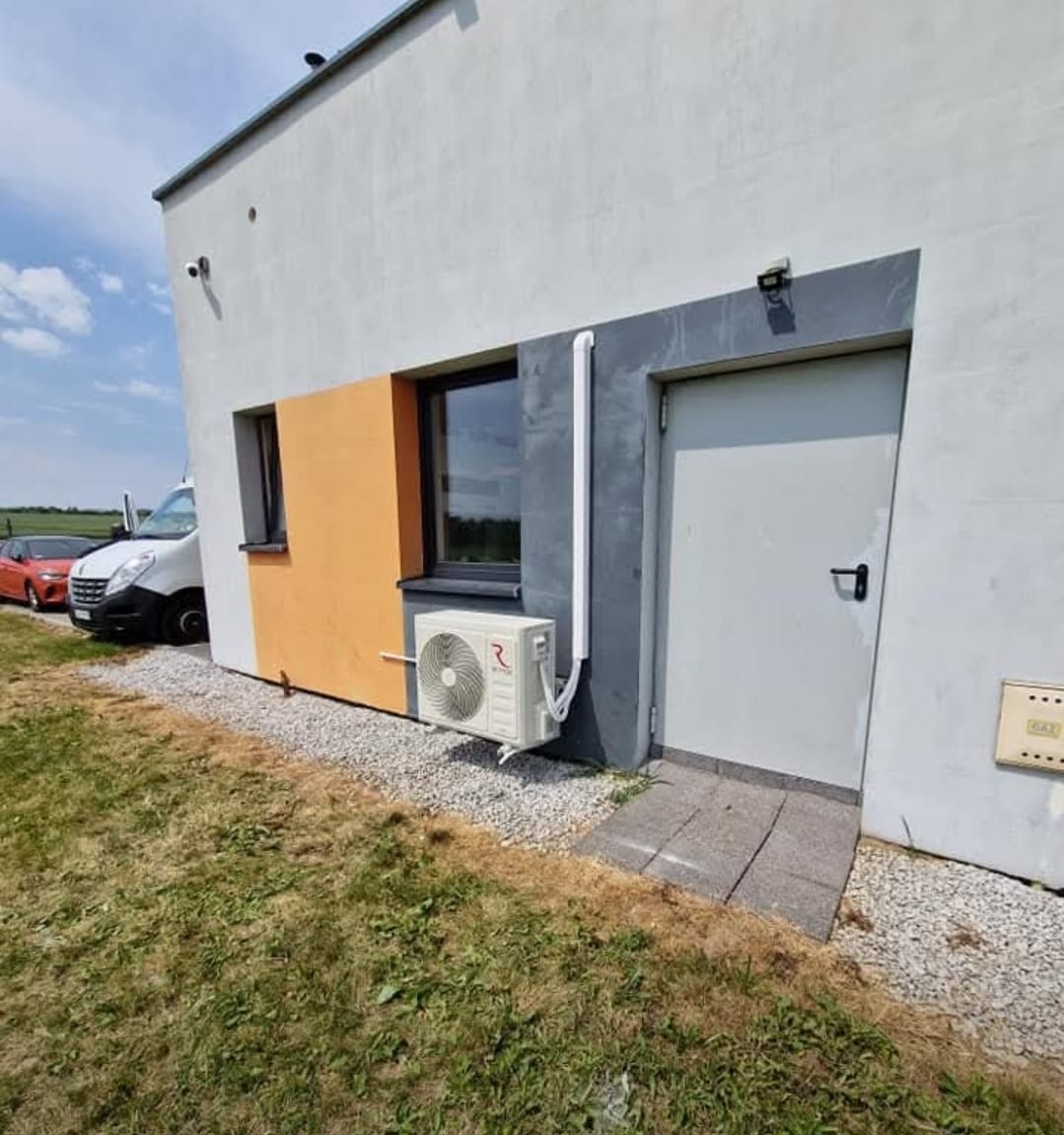 Biała klimatyzacja zewnętrzna zamontowana na elewacji budynku z szarą stolarką okienną i drzwiami. Obok zaparkowany biały bus i czerwony samochód osobowy.