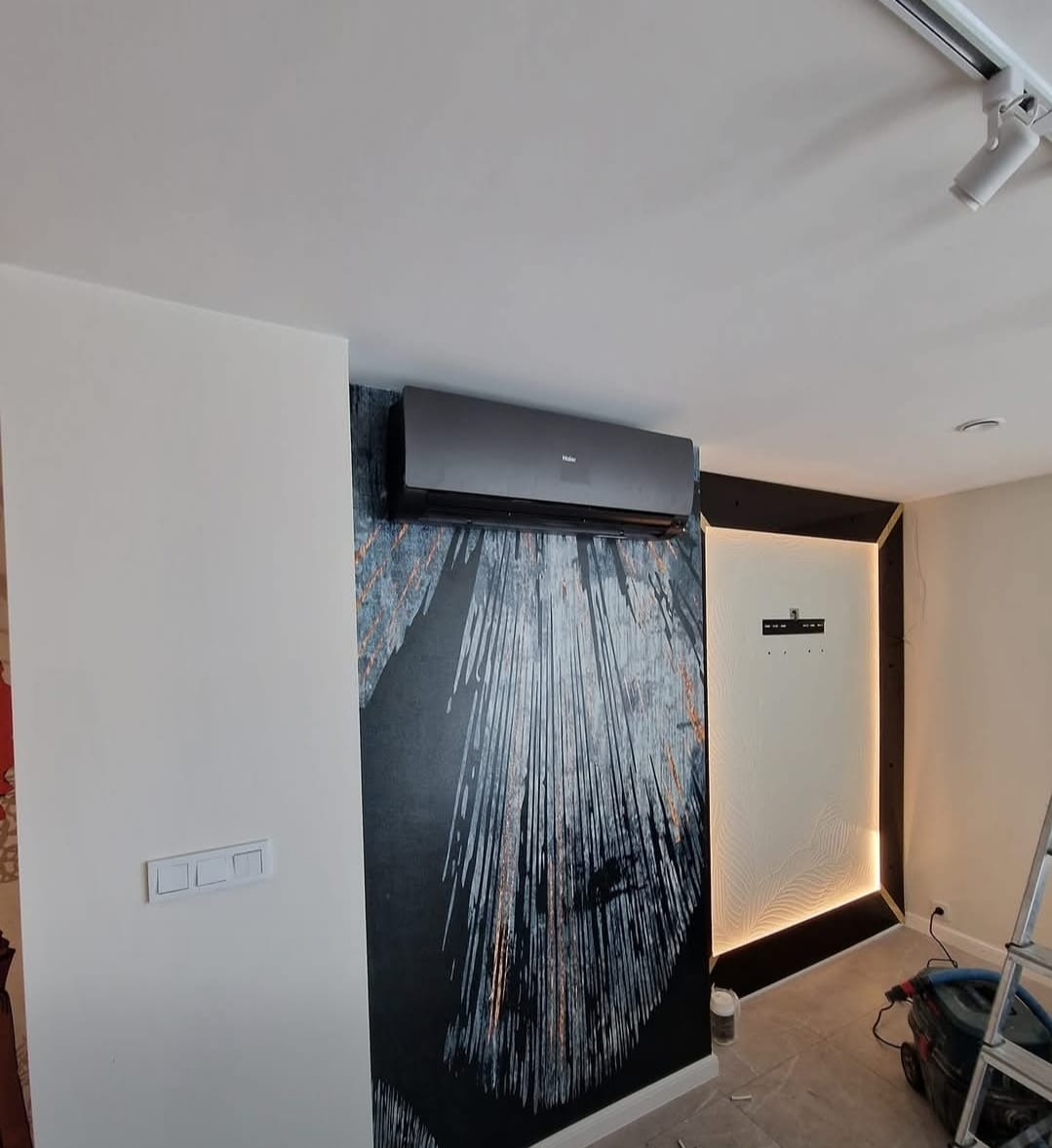 Czarny klimatyzator Haier zamontowany na ścianie z abstrakcyjną tapetą w nowoczesnym wnętrzu. Widoczny fragment drabiny i odkurzacza.
