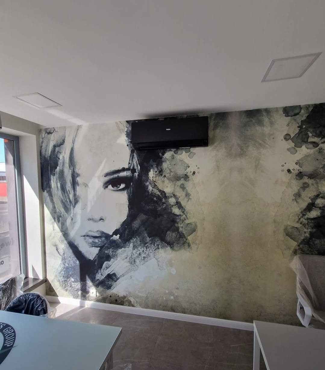 Czarny klimatyzator Haier zamontowany na ścianie z artystycznym malowidłem kobiety w odcieniach szarości i beżu. Widoczne fragmenty biura i okno.