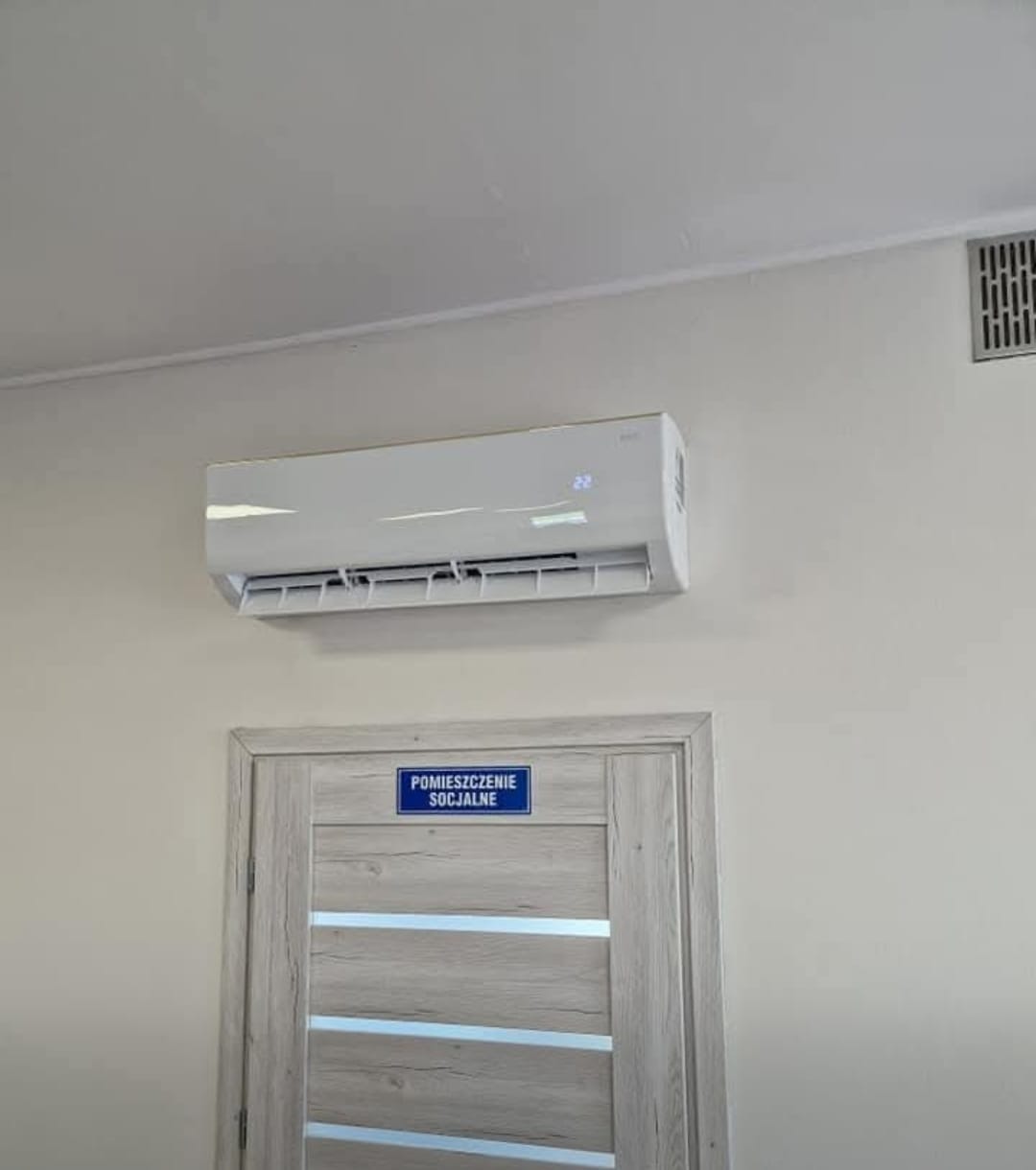 Biały klimatyzator ścienny zamontowany nad drzwiami z napisem 'Pomieszczenie Socjalne'. Widoczna kratka wentylacyjna w rogu. Ustawiona temperatura 22 stopnie.